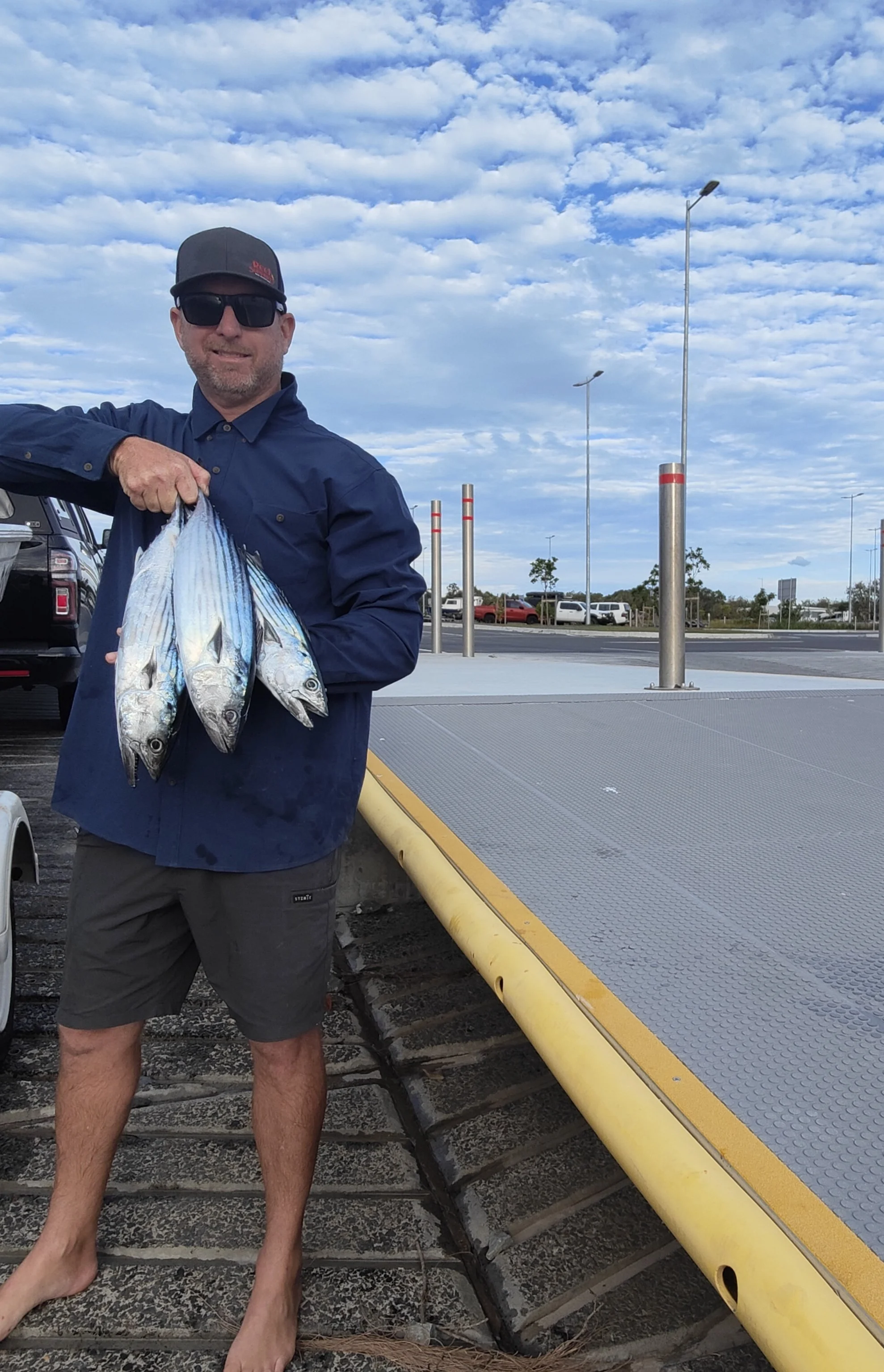 A man wearing sunglasses, a blue shirt, and gray shorts, holding several fish outdoors near a parking lot on a cloudy day.