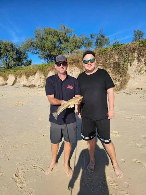 Two men standing on a sandy beach, one holding a fish, with trees and a blue sky in the background.