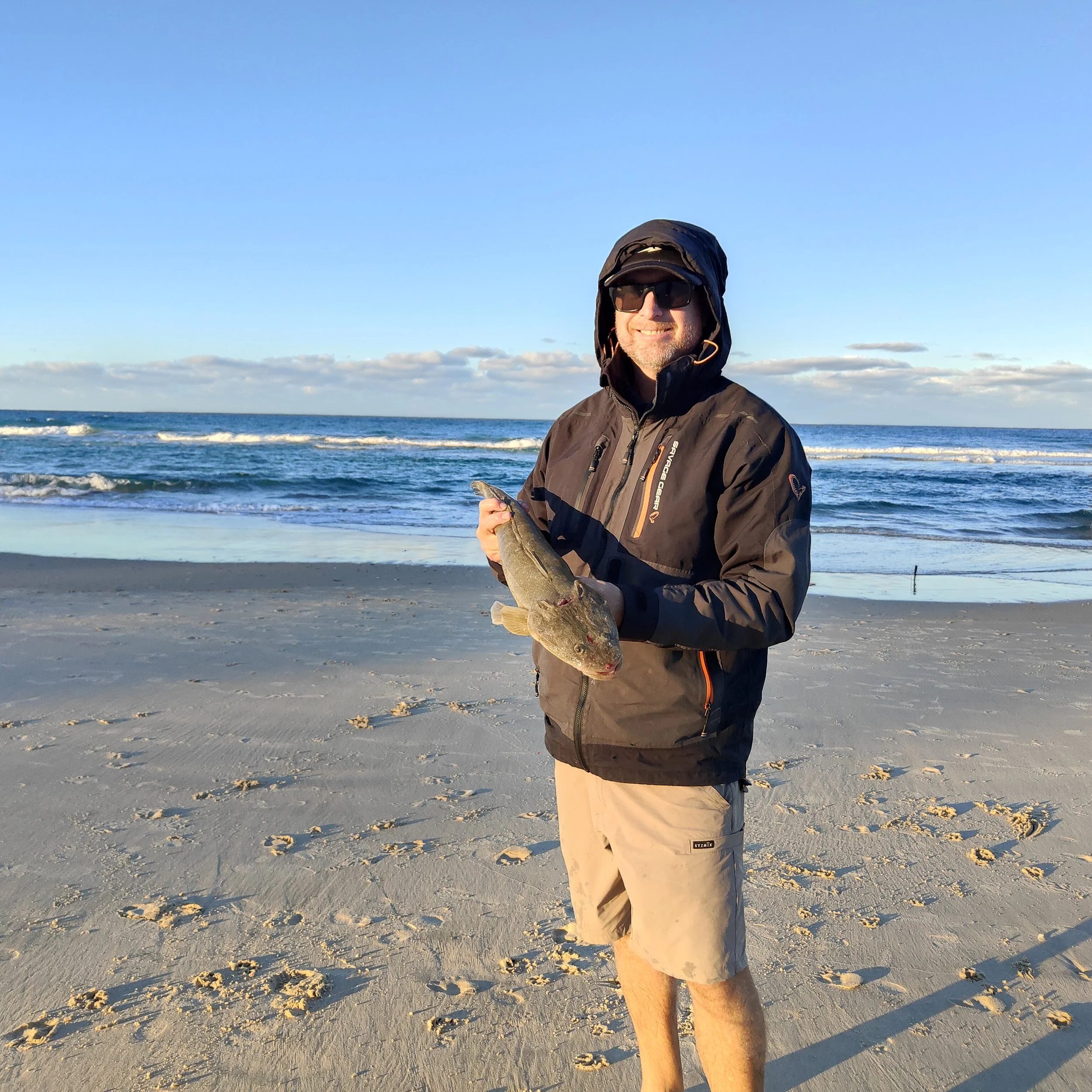 Man in black jacket, sunglasses, and khaki shorts holding a fish on a sandy beach with the ocean and sky in the background.