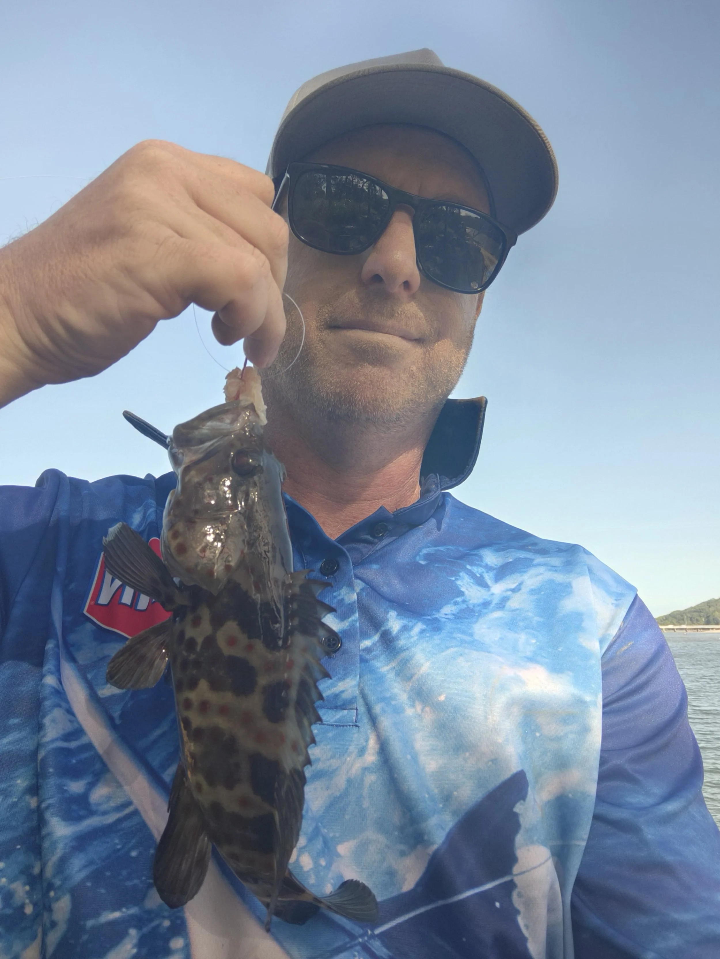Man wearing sunglasses and a cap holding a small fish, standing outdoors near a body of water under a clear blue sky.