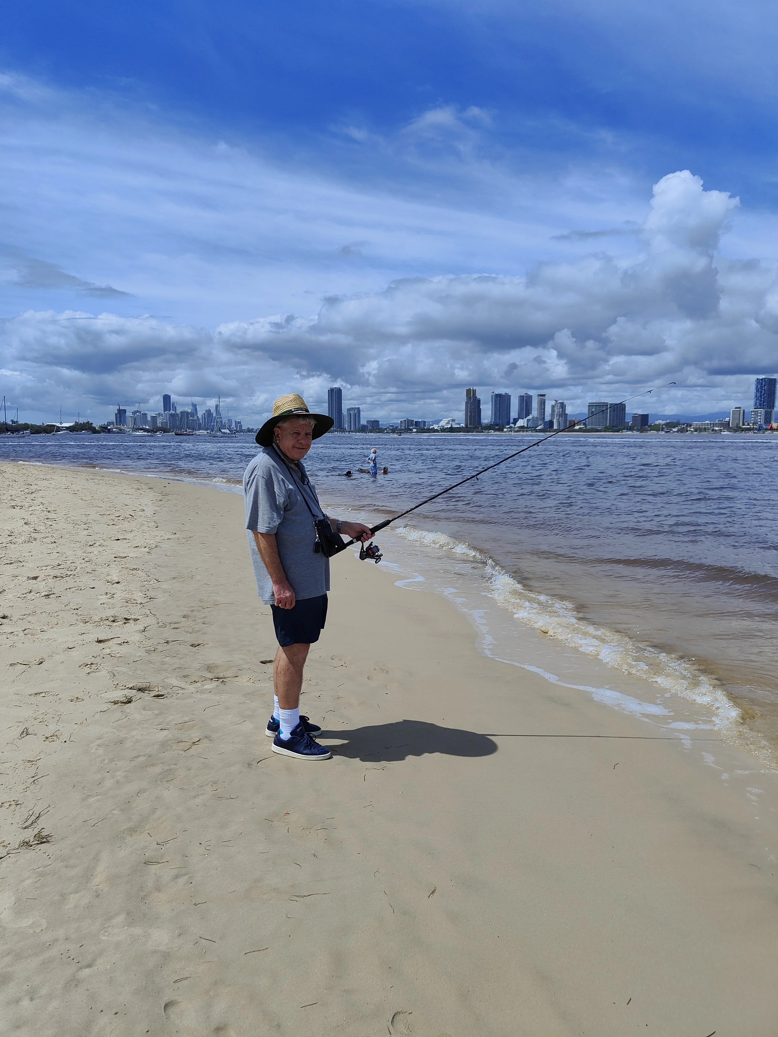 An elderly man fishing on a sandy beach with a city skyline in the background under a partly cloudy sky.