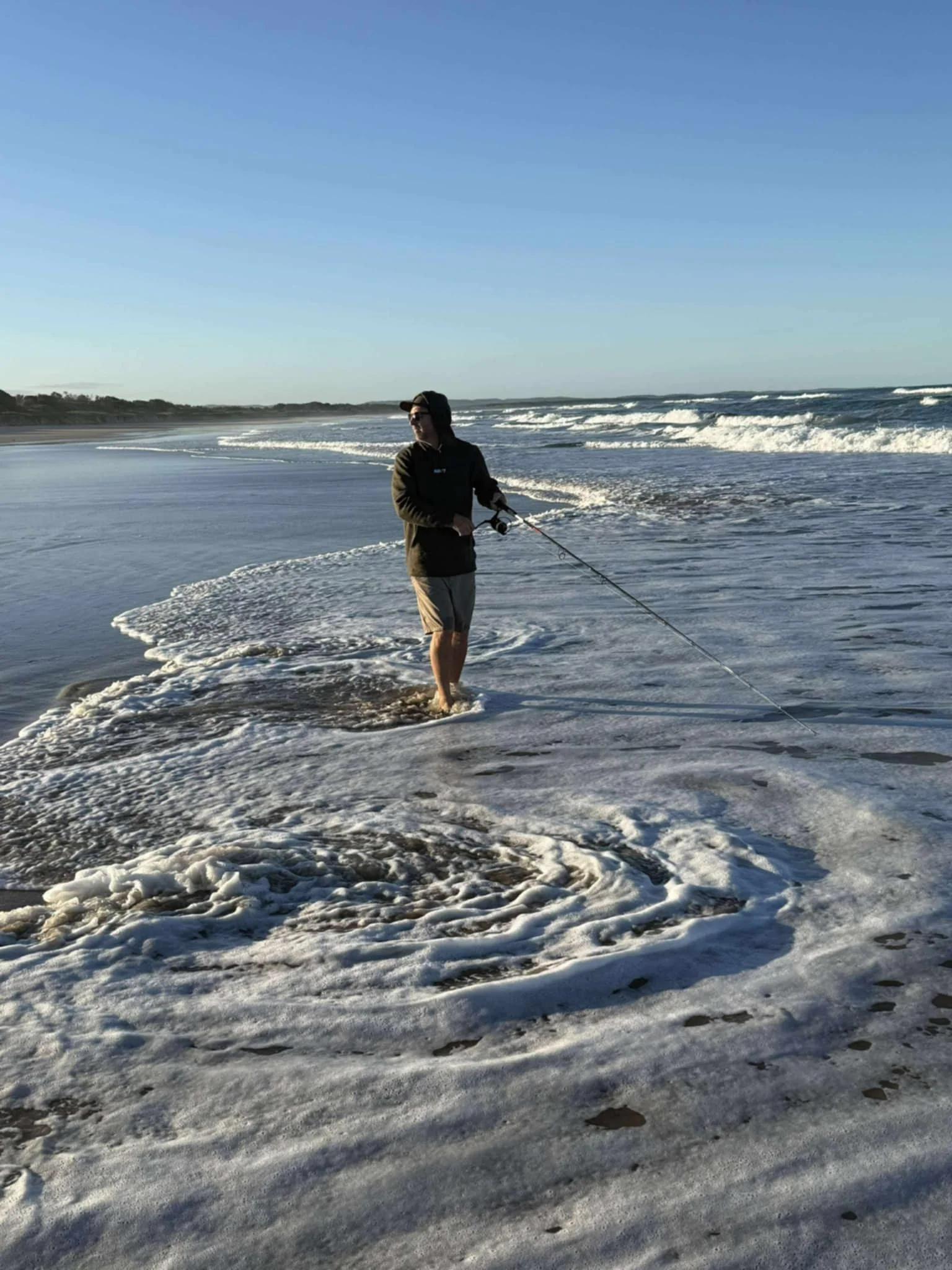 Person fishing in shallow ocean water on a sandy beach under a clear blue sky.
