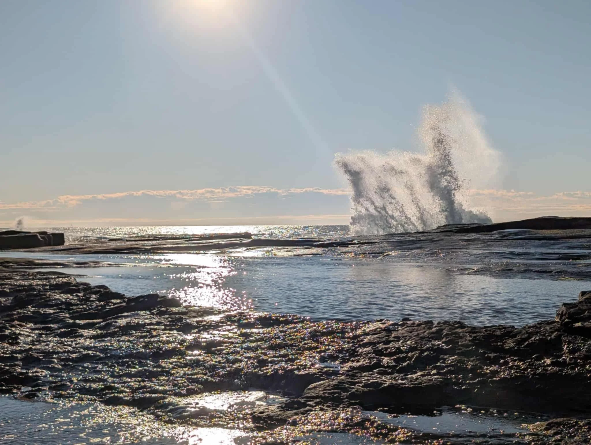 Ocean waves crashing on rocks with sunlight reflecting on the water under a clear sky.