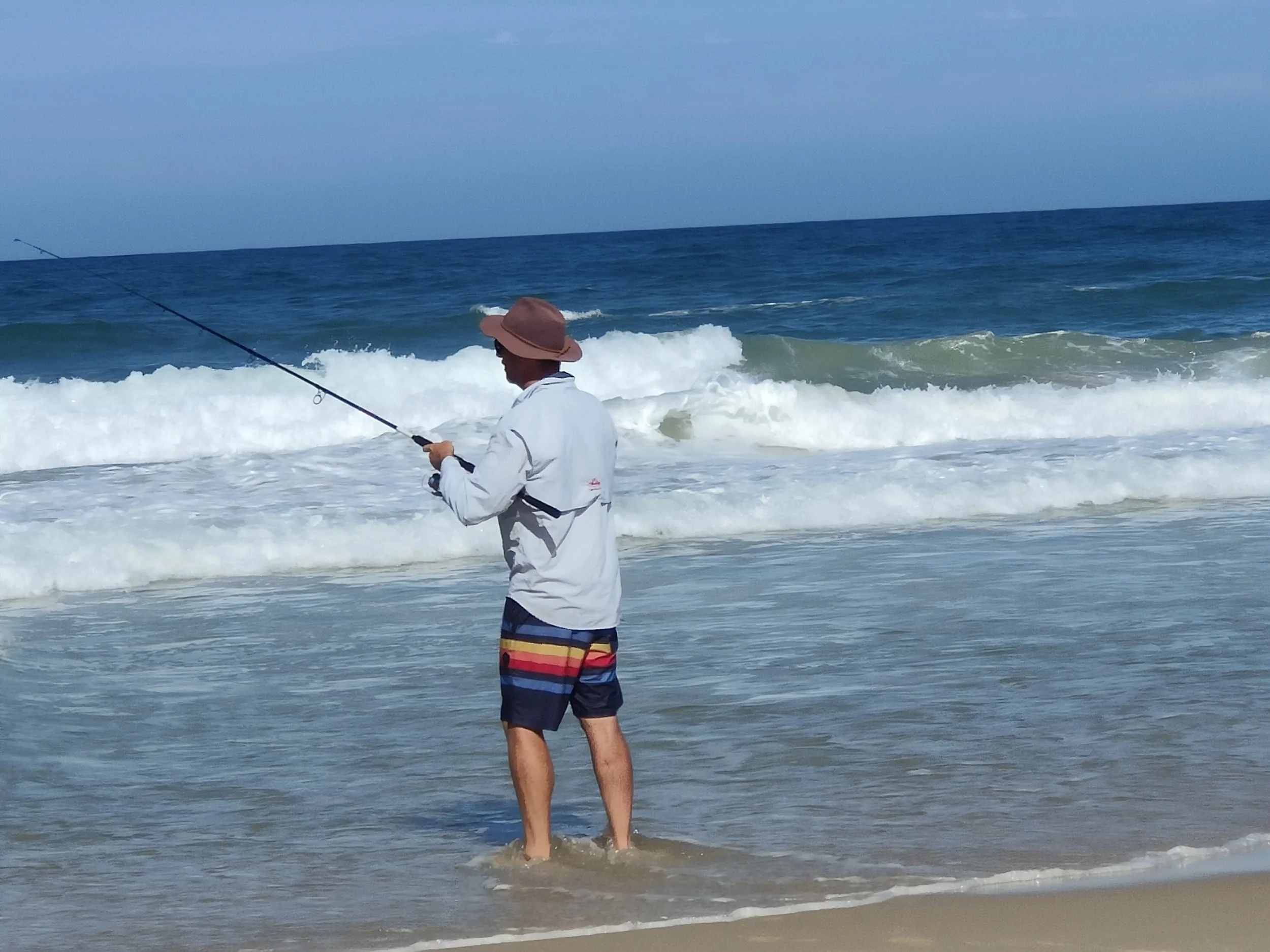 A man standing in shallow water on a beach, fishing with a rod and reel, wearing a hat, long sleeve shirt, and colorful shorts, with waves crashing behind him and the ocean extending to the horizon.