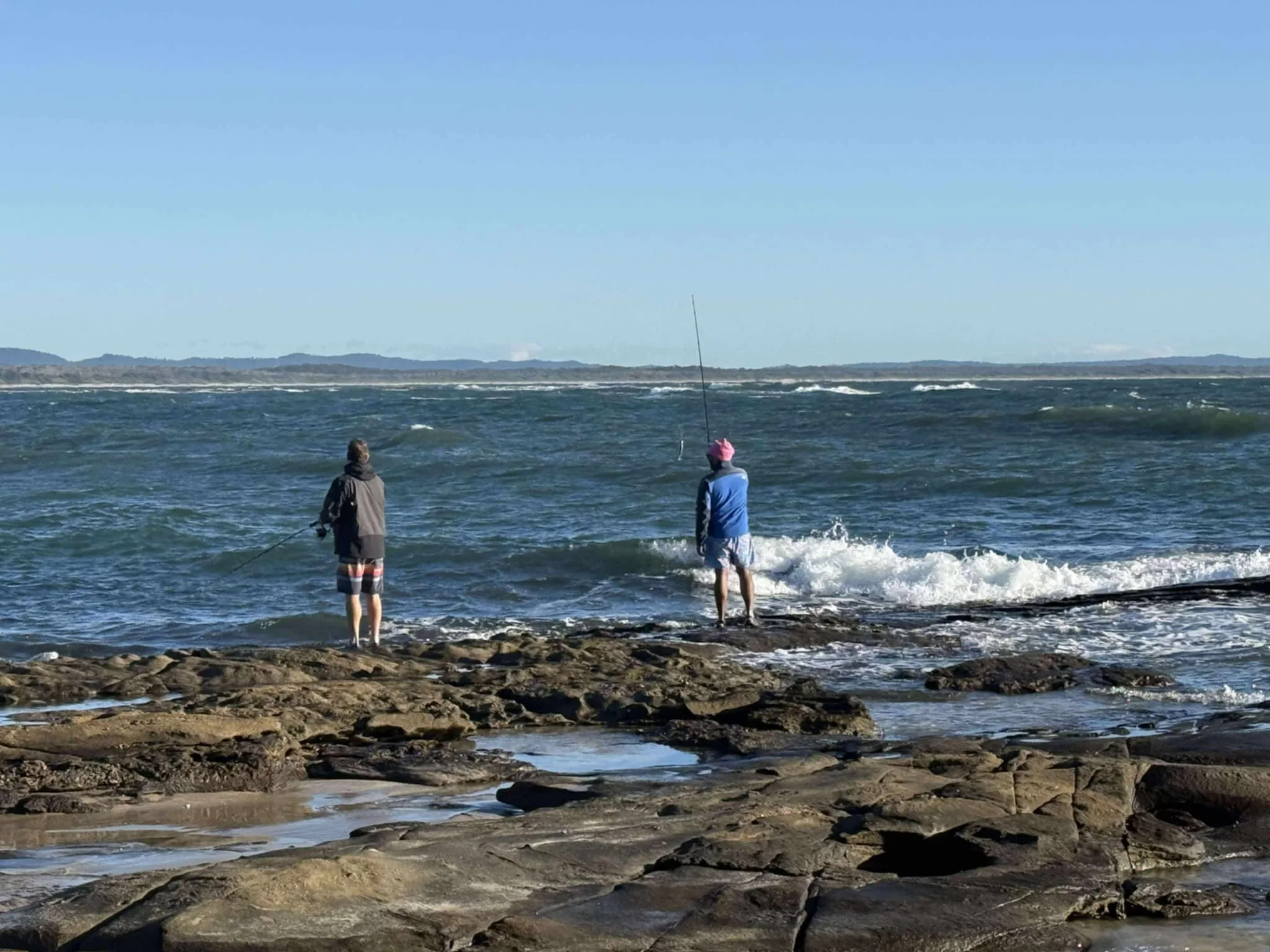 Two people fishing on a rocky beach near the ocean with waves and a distant shoreline under a clear blue sky.