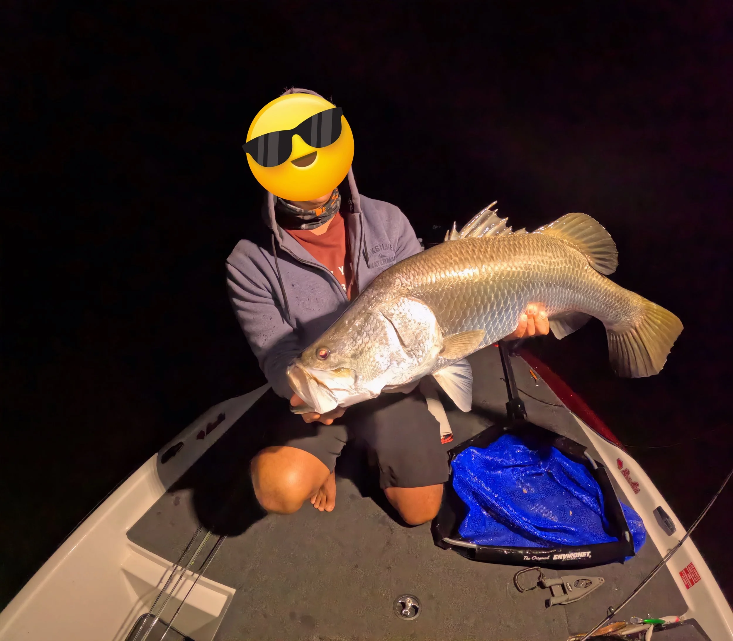 Person on boat holding a large fish at night with a fun emoji face wearing sunglasses covering their face.