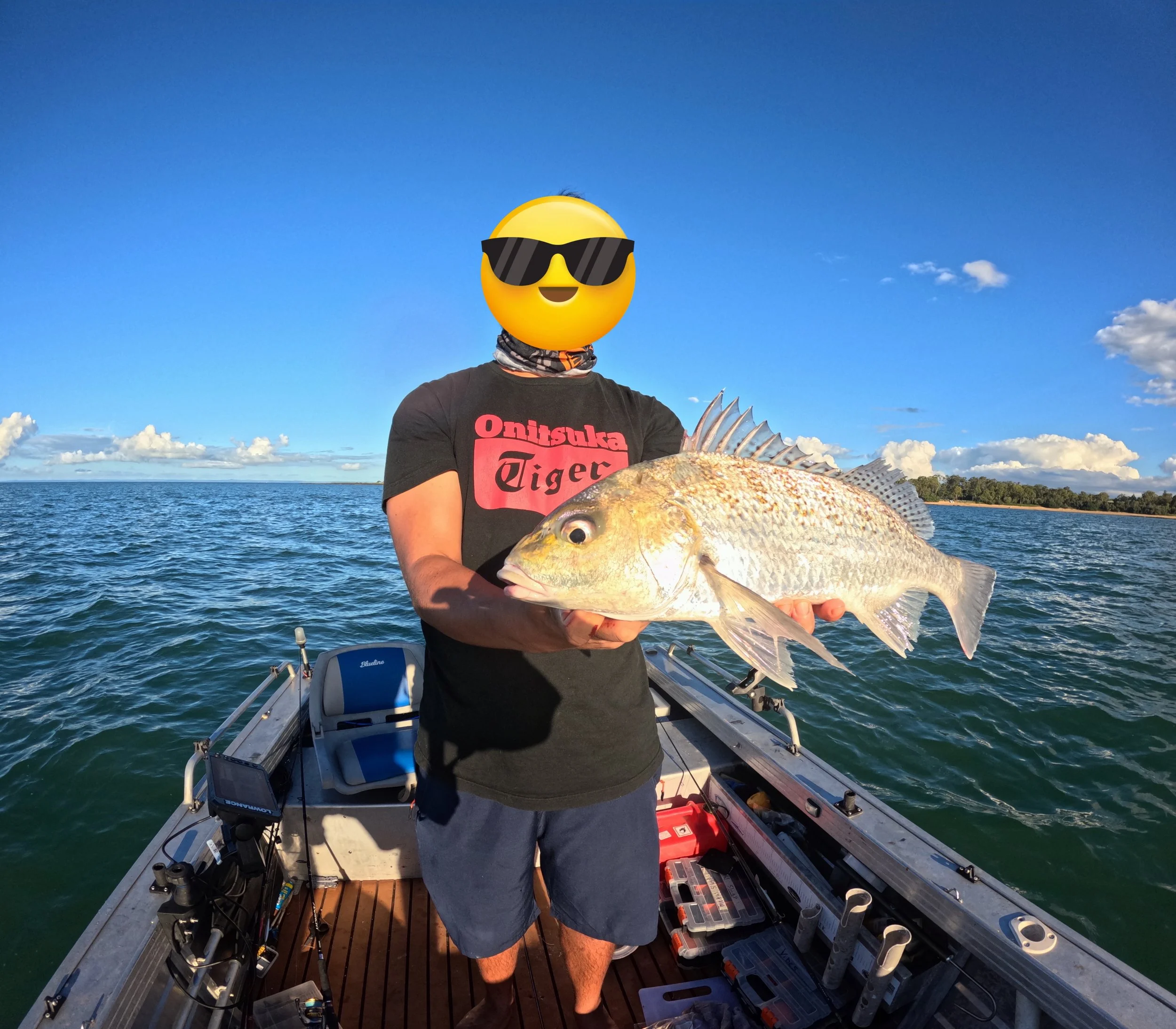 Person on boat holding a large fish, wearing sunglasses emoji face, black T-shirt with red text, blue shorts, with water and sky background.