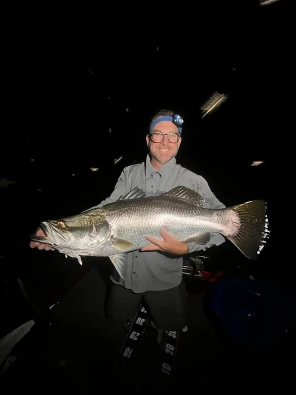 A man wearing glasses, a headlamp, and a light gray shirt is smiling and holding a large fish horizontally in a dark setting.