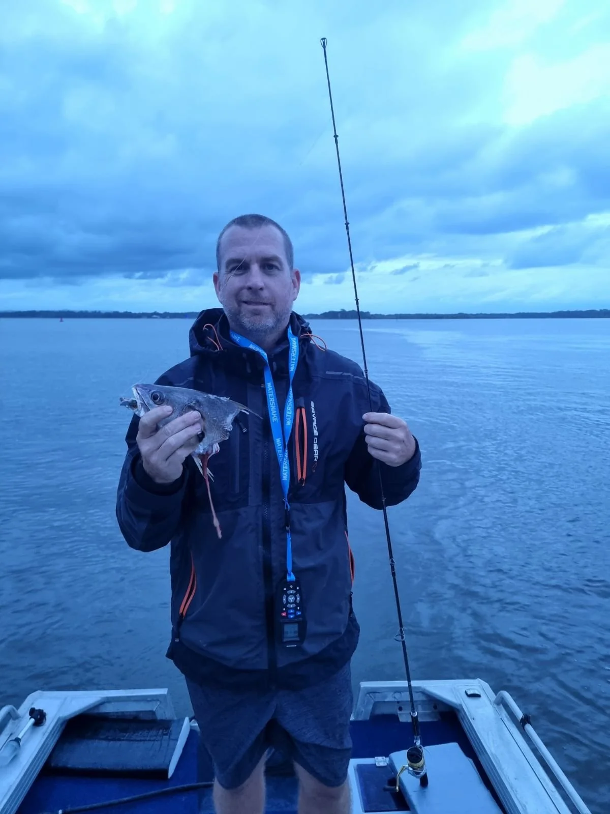 Man holding a fish on a boat with a fishing rod, on a calm body of water under cloudy sky.