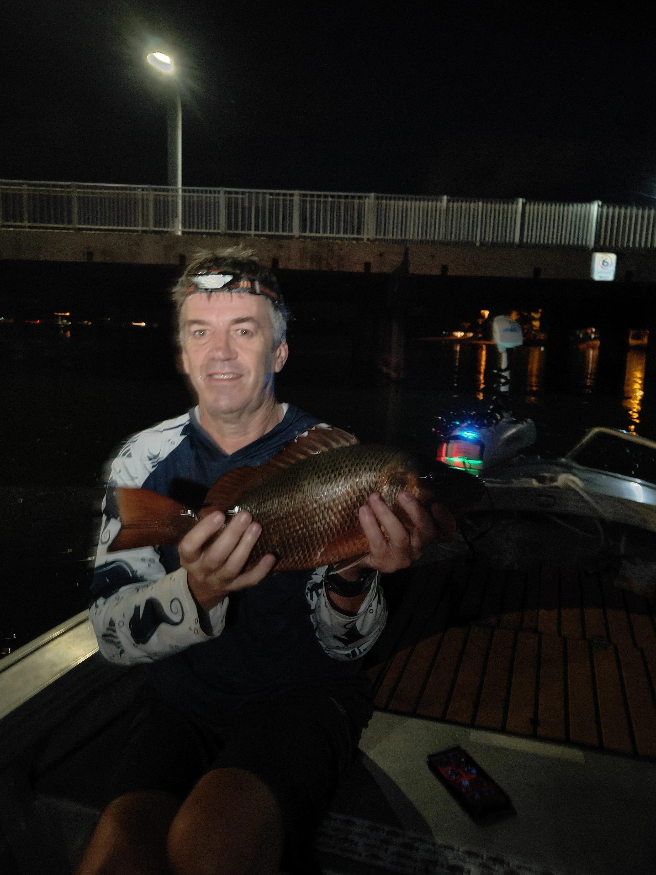 A man holding a large fish on a boat at night, with a bridge and water in the background.