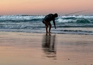 Person fishing on the beach at sunset with waves in the background.