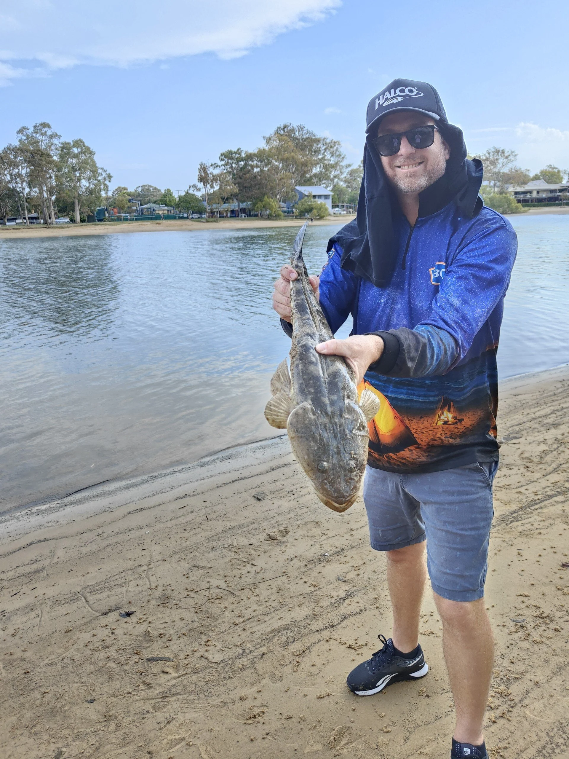 A man holding a fish near a body of water on a sandy beach, wearing sunglasses, a cap, a blue jacket, shorts, and sneakers.