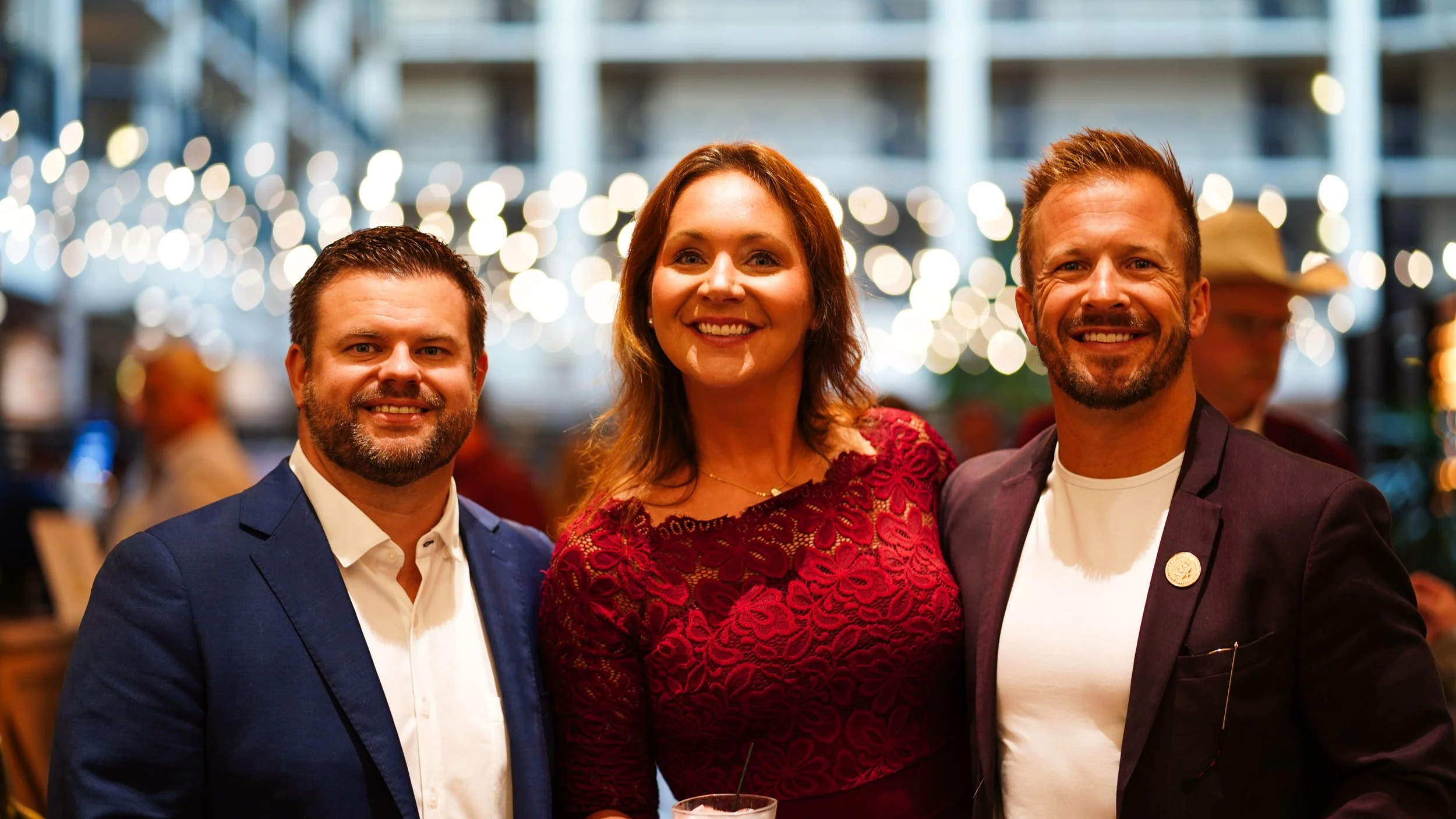 Three smiling people at a social event with string lights in the background. Two men are wearing suits, and a woman in a red dress is in the middle.