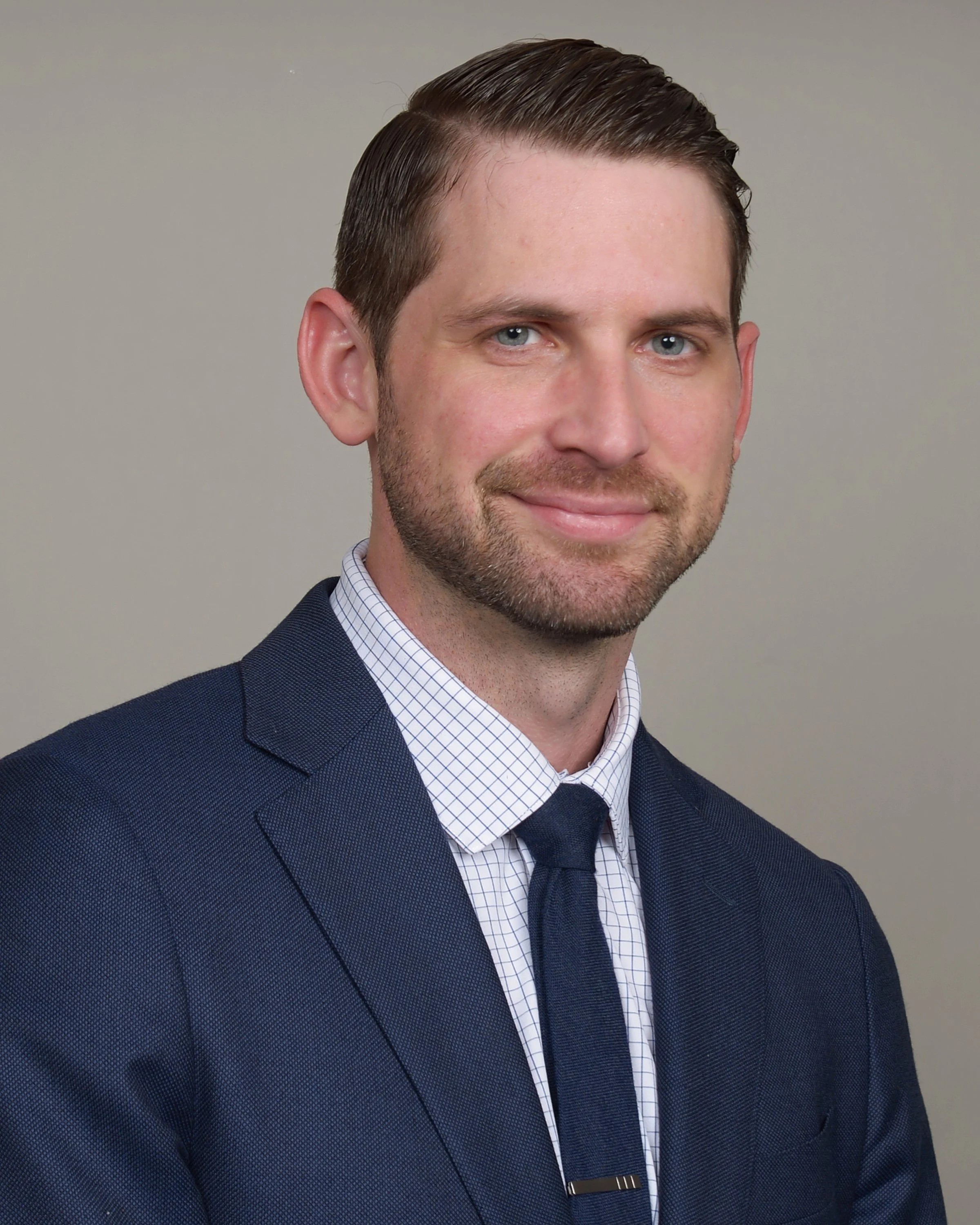 Professional headshot of a man with short brown hair, blue eyes, and a light beard, wearing a navy suit, white checkered shirt, and a navy tie, smiling slightly, against a plain gray background.