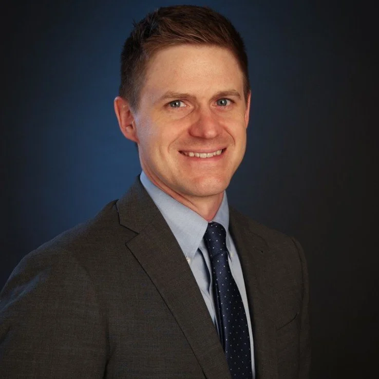A man in a dark suit, light blue shirt, and navy tie smiling against a dark background.