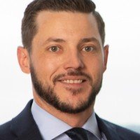 A man with dark hair and a beard wearing a suit and smiling.