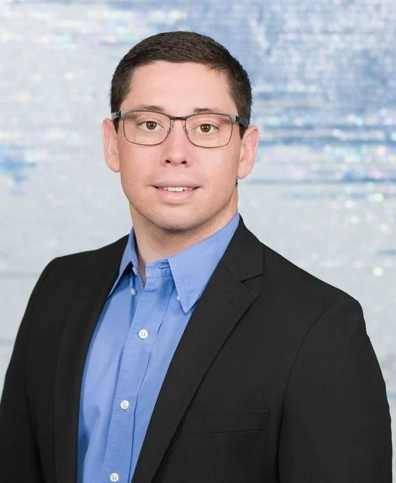 Professional headshot of a man wearing glasses, a blue shirt, and a black blazer, standing in front of a blurred blue and white background.