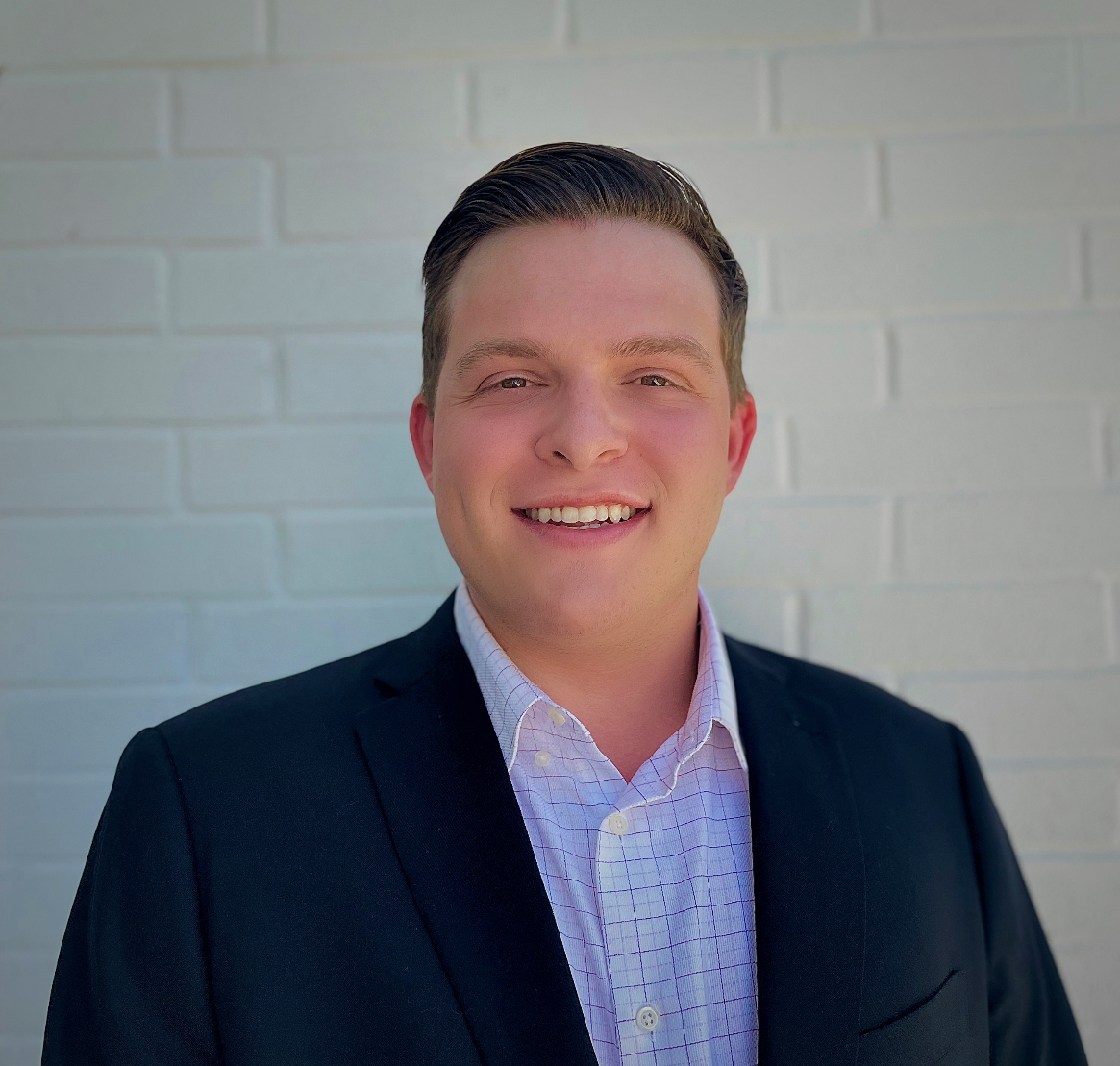 A smiling young man in a dark suit jacket and white checkered shirt standing in front of a white brick wall.