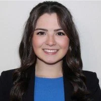 A young woman with long dark hair, smiling, wearing a black blazer and a blue top, against a plain background.