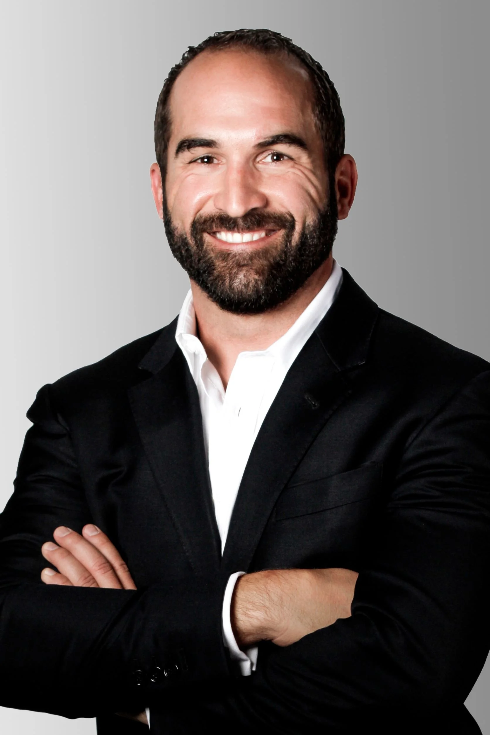 A smiling man with a beard, wearing a black suit and white shirt, standing with crossed arms against a gray background.