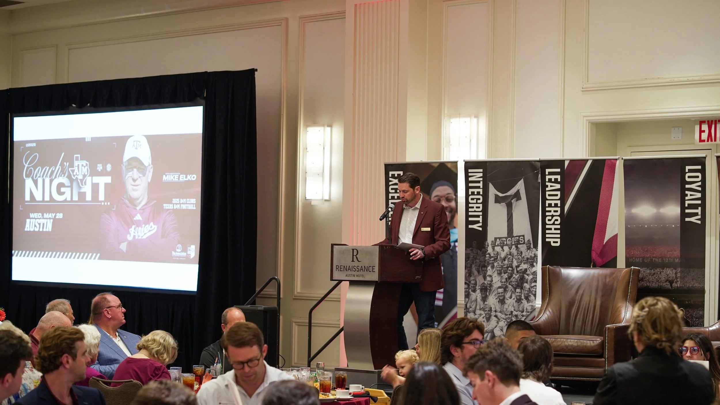Man in a maroon blazer and white shirt speaking at a podium during a formal event at the Renaissance Austin Hotel, with banners displaying words like 'Integrity,' 'Leadership,' and 'Loyalty,' and an audience seated at round tables.