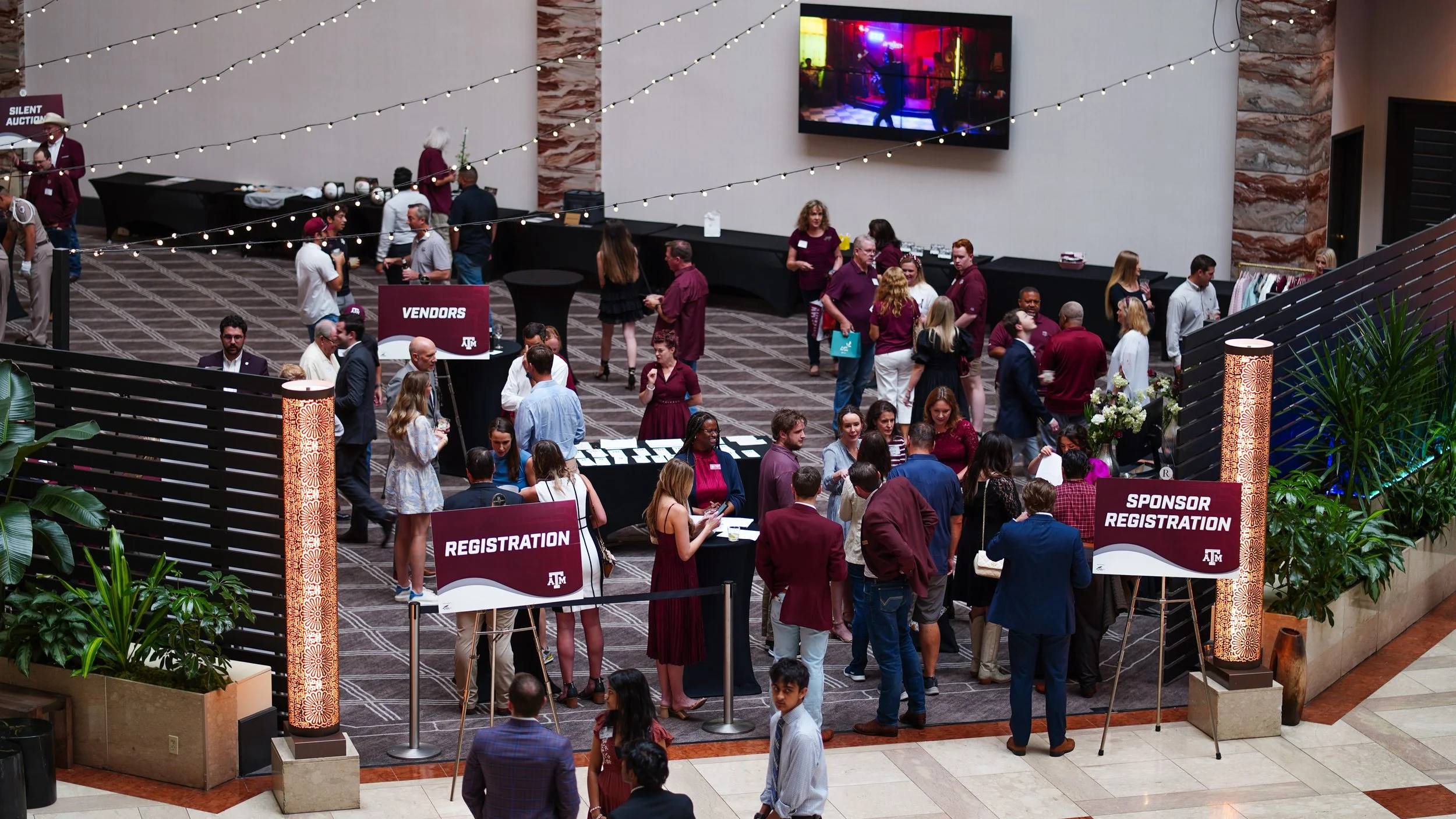 People attending a conference registration area at a hotel or convention center, with signs for 'Registration,' 'Vendors,' and 'Sponsor Registration'; some people are talking, walking, or registering at tables, with decorative lighting and plants in 
