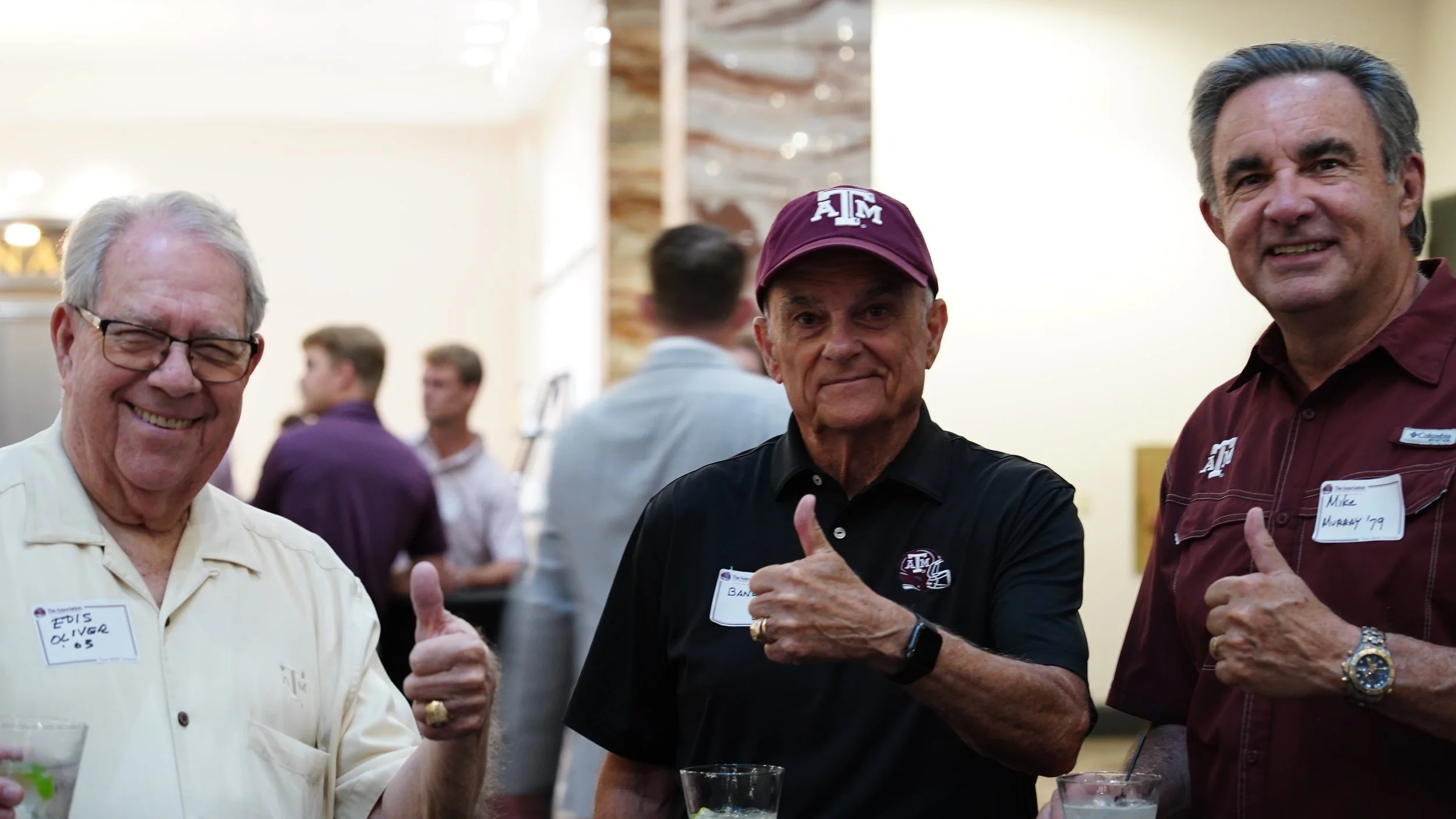 Three men at a social event, giving thumbs-up, smiling, wearing name tags, two with Texas A&M clothing, one with eyeglasses and a watch, background with other guests.