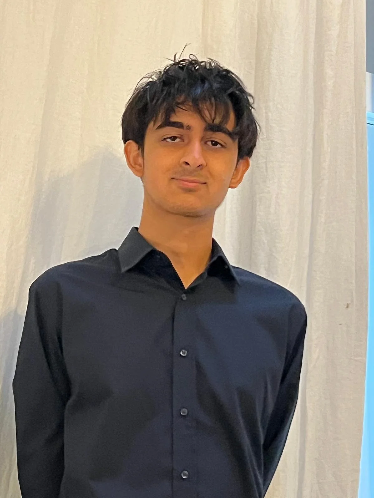 Young man with black hair, wearing a black dress shirt, standing in front of a light-colored curtain.
