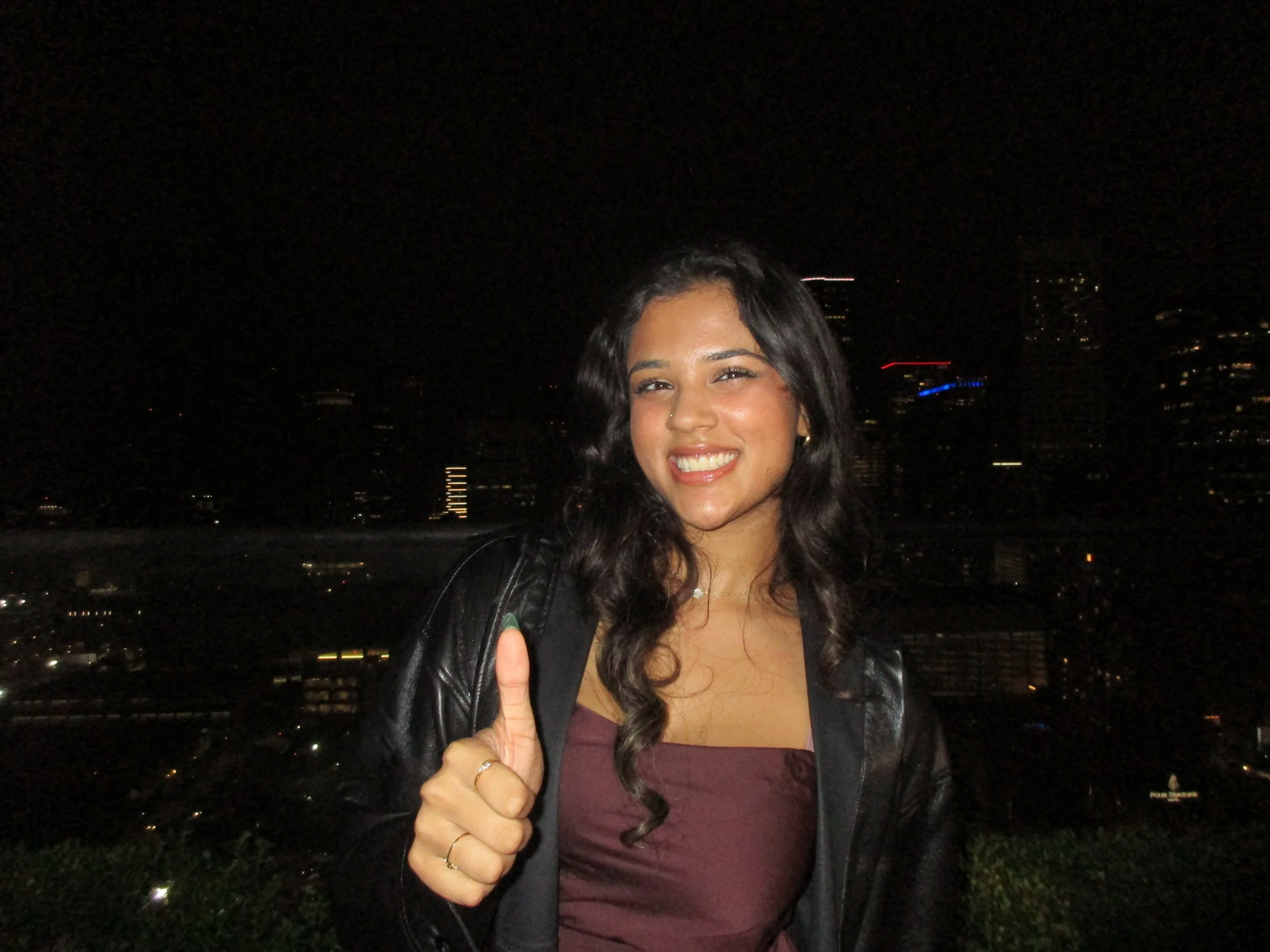 Young woman smiling at night, giving a thumbs-up gesture, city skyline in background.