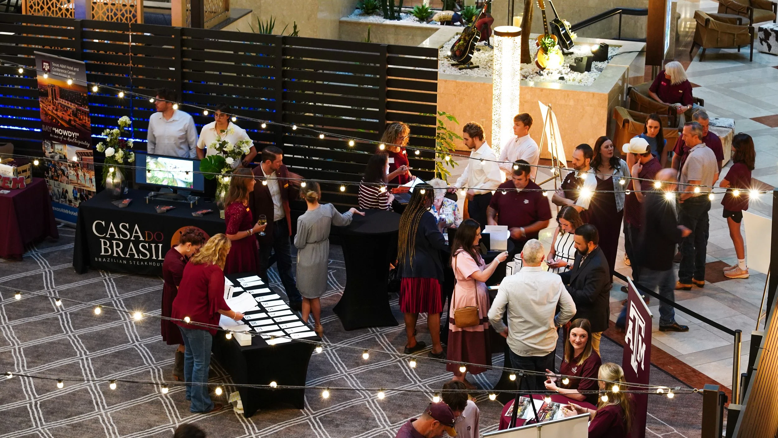 A busy indoor event or conference with multiple booths, including a Brazilian restaurant, and attendees engaging, networking, and browsing. String lights and decor adorn the space.
