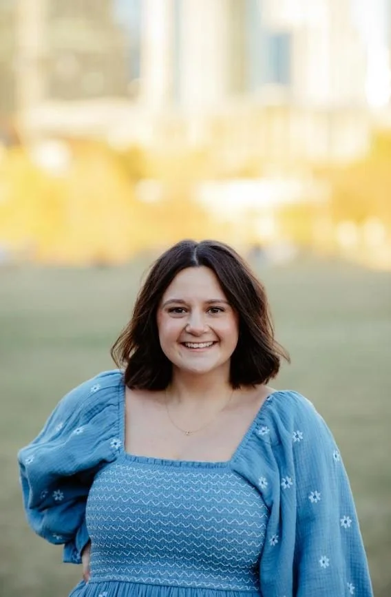 A woman with shoulder-length brown hair smiling outdoors in a blue dress with puffed sleeves and embroidery, with a blurred background of a grassy area and buildings.