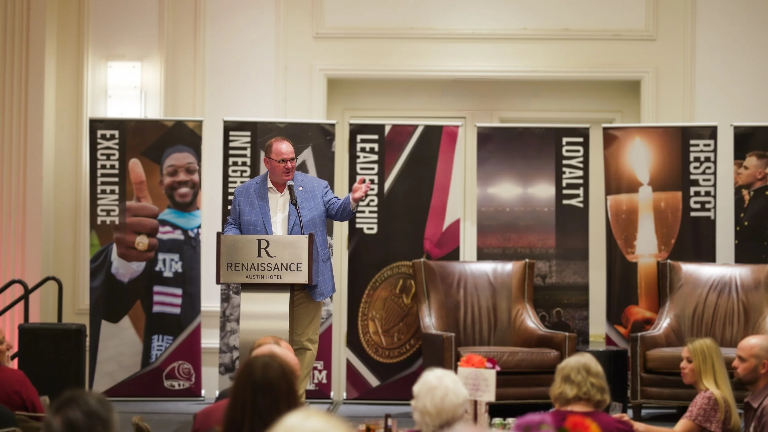 A man in a light blue plaid blazer giving a speech at a podium during an event at the Renaissance Austin Hotel. Behind him are vertical banners with words like 'Excellence,' 'Integrity,' 'Leadership,' 'Loyalty,' and 'Respect,' and images including a smiling graduate with a thumbs-up, a medal, a candle, and a military officer. The audience is seated and listening attentively.