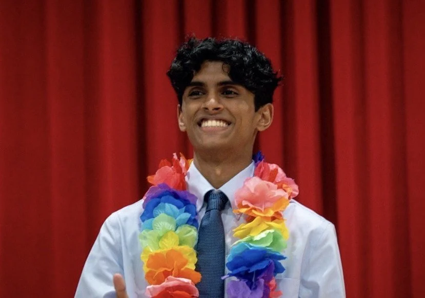 Young man smiling with a colorful lei and formal attire, standing in front of red curtains.