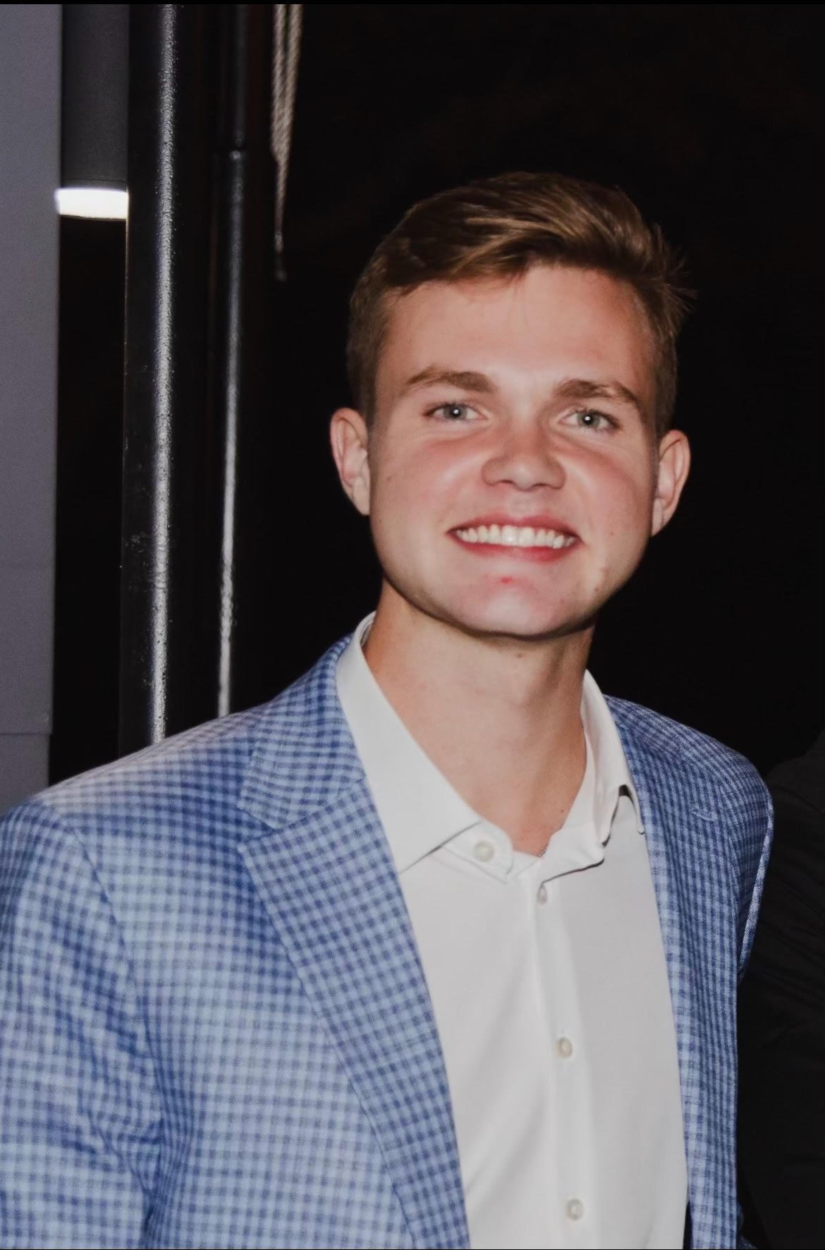 A young man with short brown hair, blue eyes, wearing a white collared shirt and a blue plaid blazer, smiling at the camera.