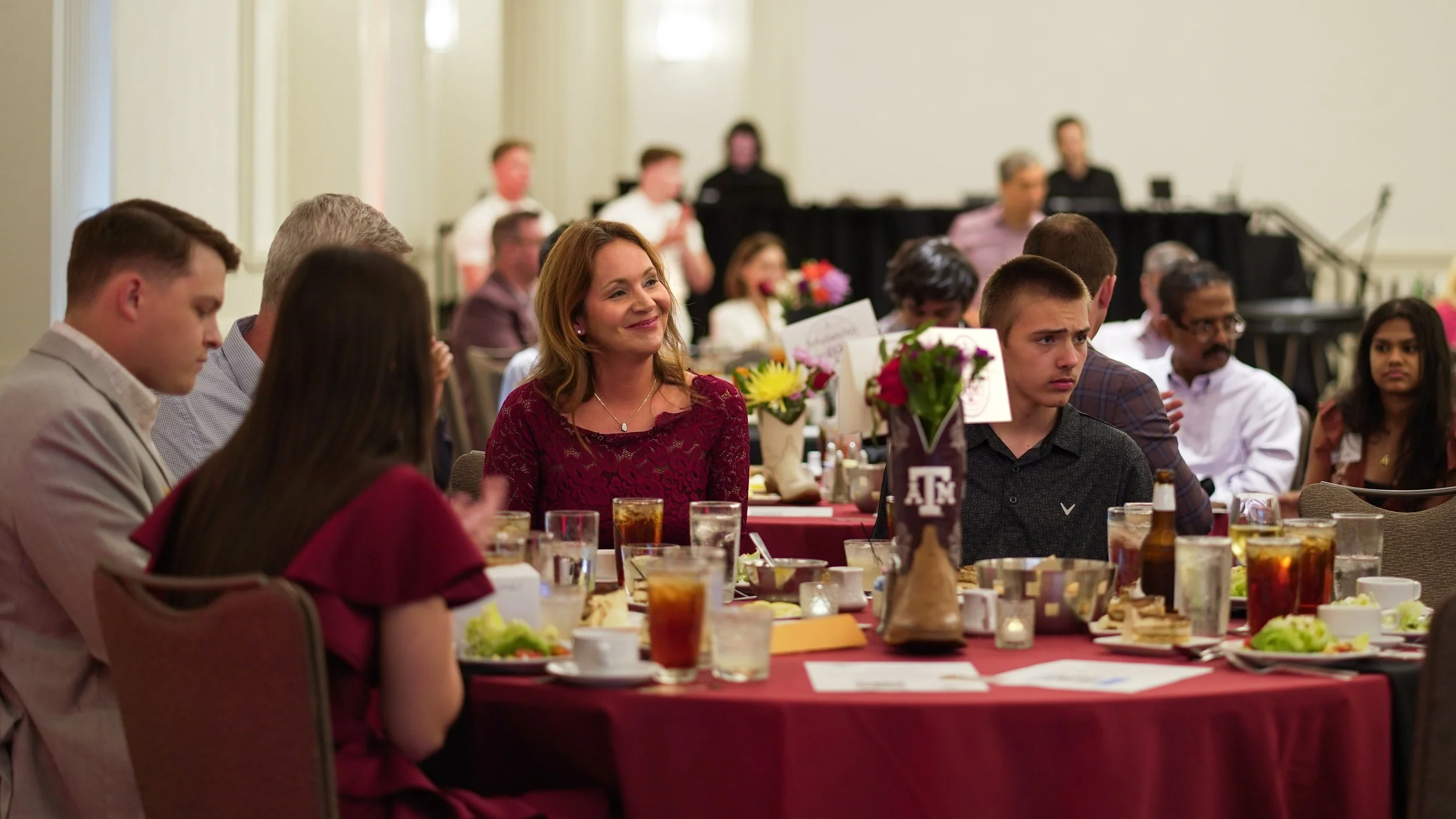People seated at a dining table during a formal event, with a woman smiling in the center and floral decorations on the table.