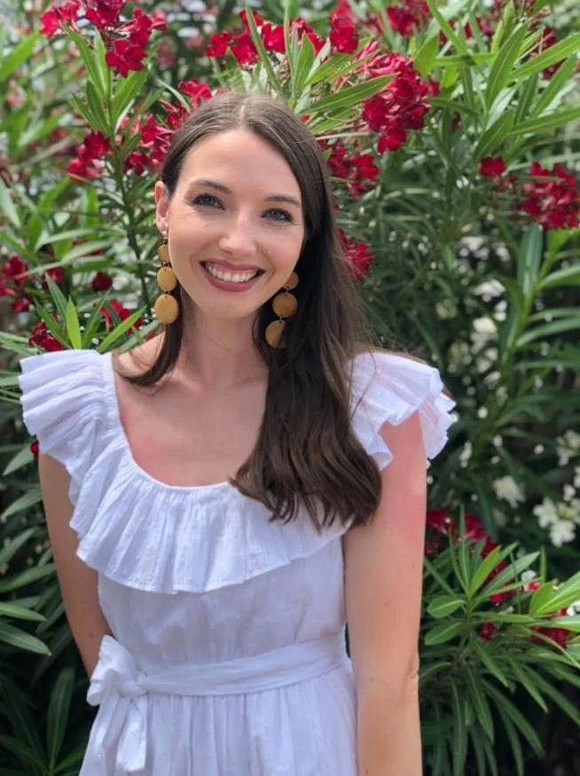 A woman with long brown hair, wearing a white ruffled dress and large yellow earrings, smiling in front of red and white flowers and green foliage.