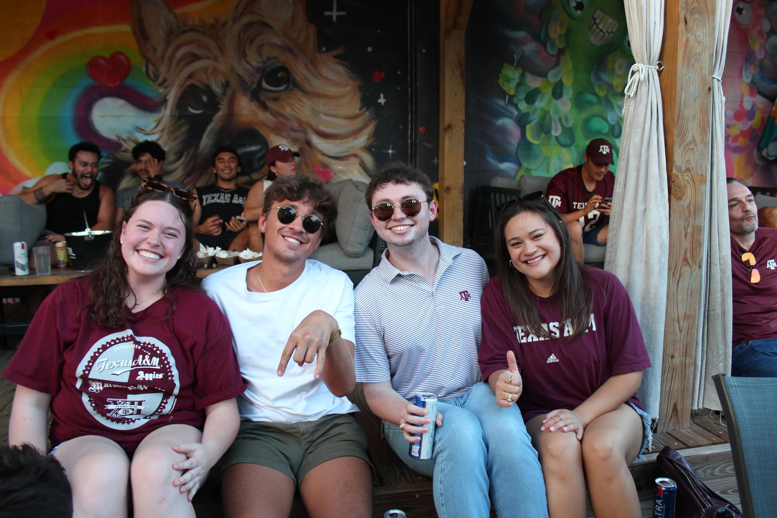 Group of four students sitting together outdoors, smiling, some wearing University of Texas A&M apparel, with a colorful mural of a lion and rainbow in the background, and other people in Texas A&M shirts in the background.
