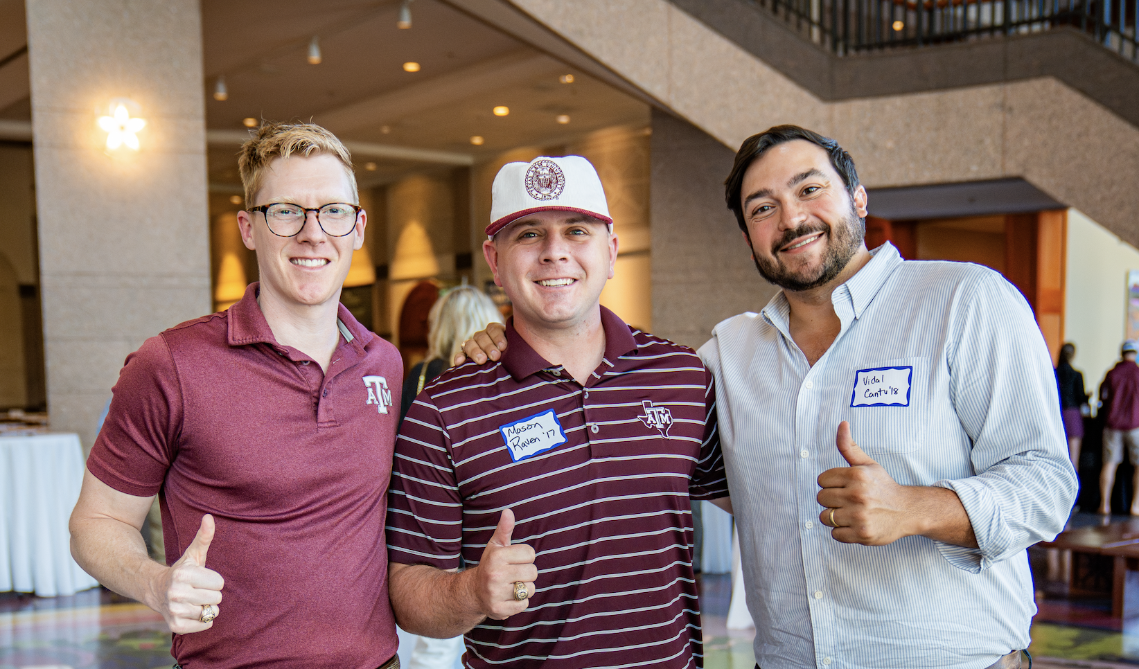 Three men standing together at an indoor event, smiling and giving a thumbs-up. They are wearing casual and company-branded clothing, with name tags on their shirts. The background shows a lobby or reception area with other people and warm lighting.