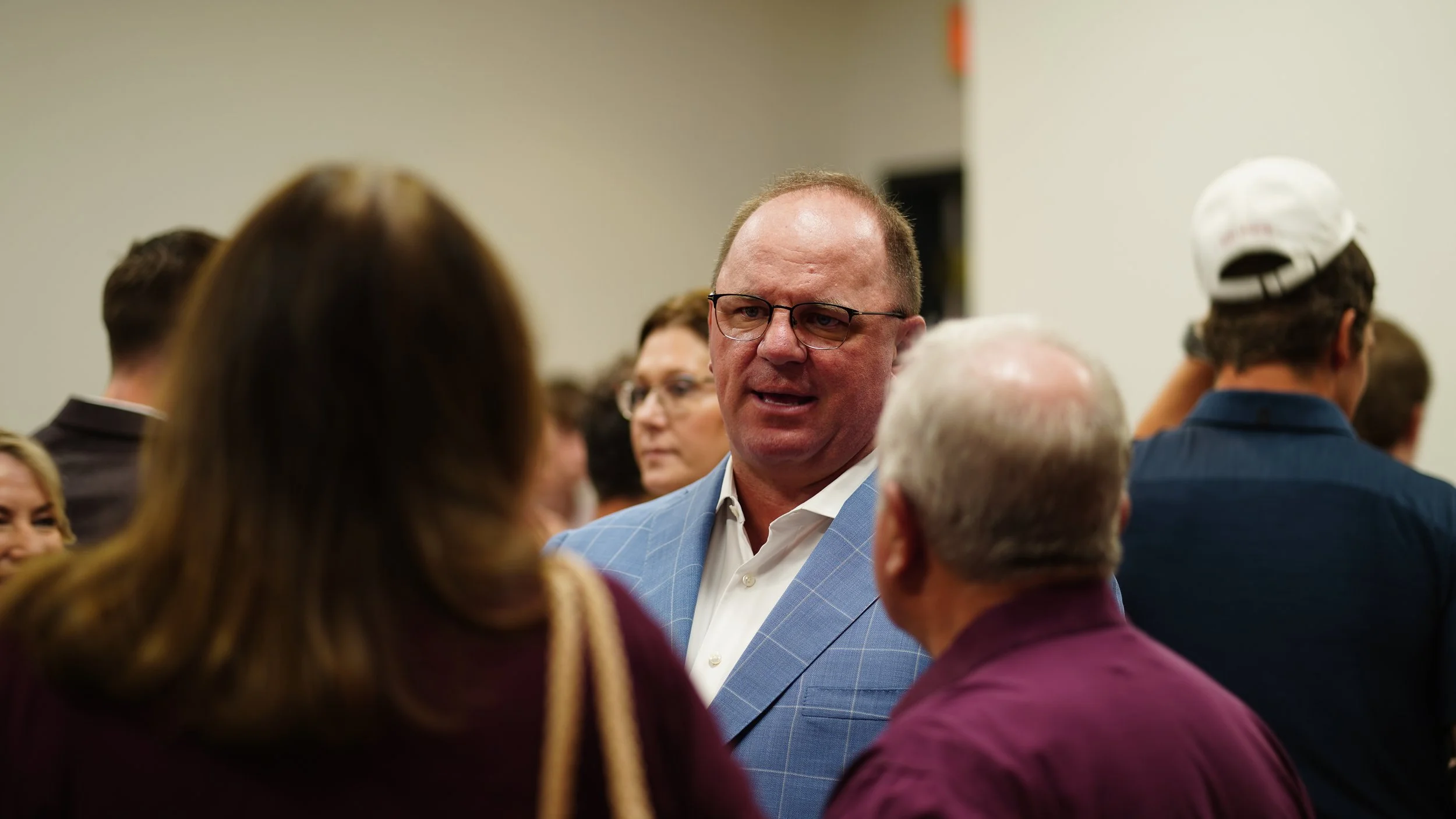 A man in a light blue suit and glasses is talking to a group of people at a social event or conference.