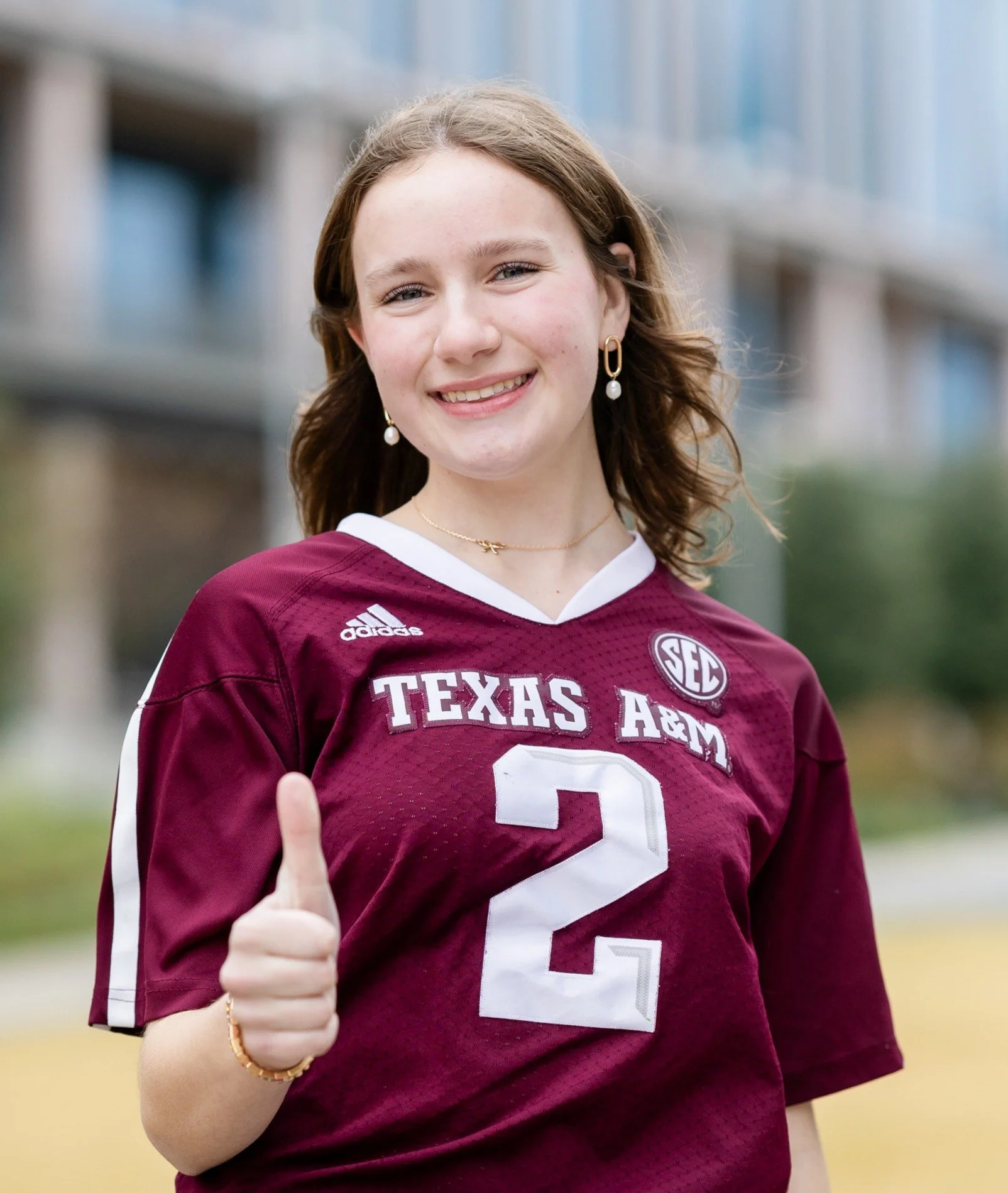 A smiling young woman wearing a maroon Texas A&M football jersey with the number 2, giving a thumbs-up gesture outside.