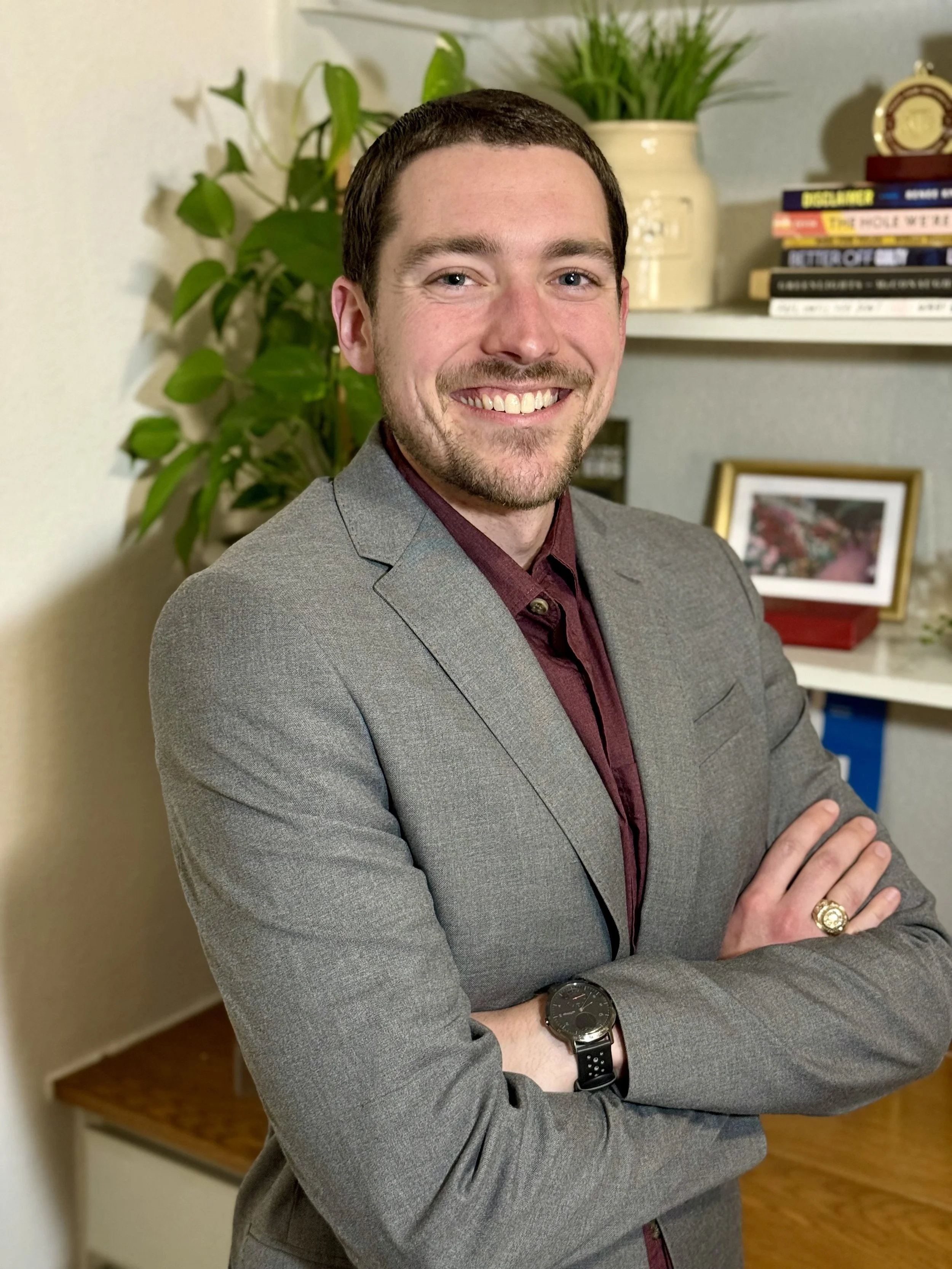 A smiling man with dark hair, wearing a gray blazer and maroon shirt, standing with arms crossed in a room with a plant, a bookshelf, and a framed photo on a shelf behind him.