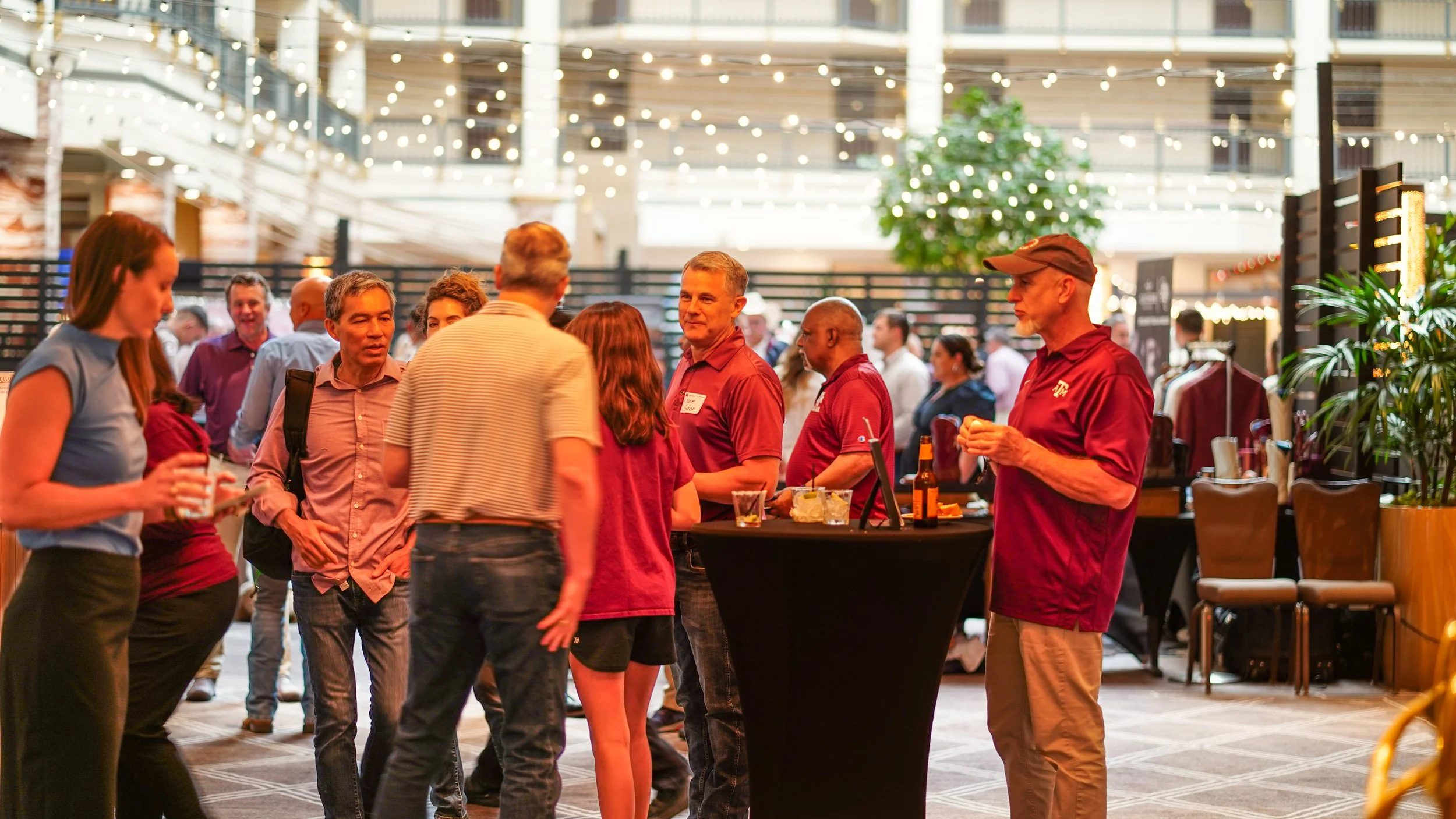 Group of people socializing at a networking event in a well-lit indoor space decorated with string lights, potted plants, and display tables.
