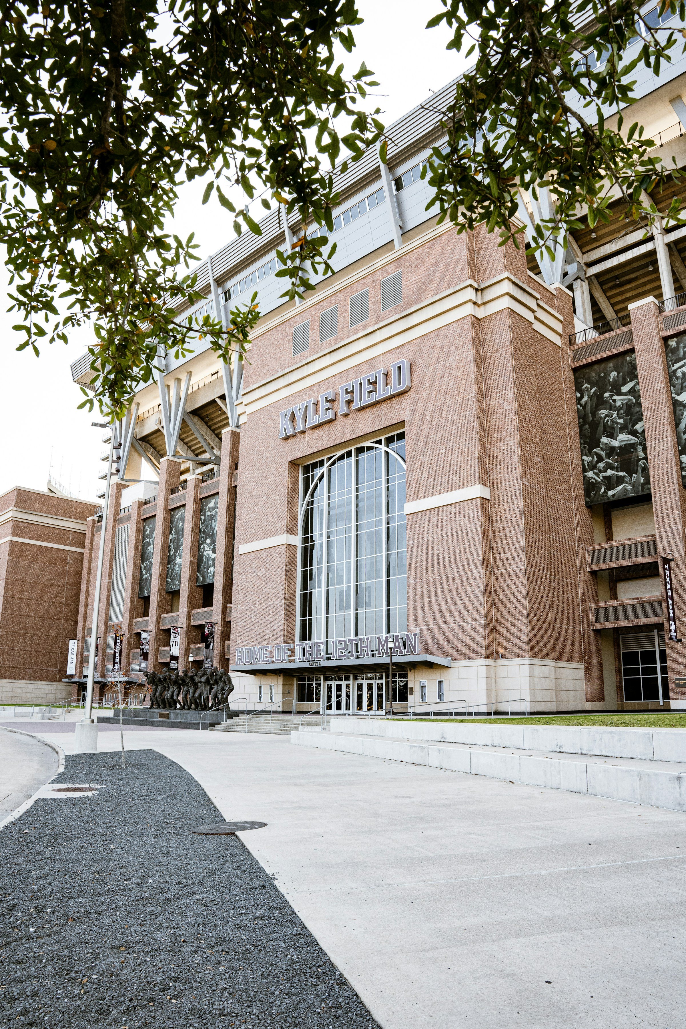 Exterior view of Kyle Field stadium with the sign reading 'Kyle Field' and 'Home of the 12th Man,' showing the entrance and some trees in the foreground.