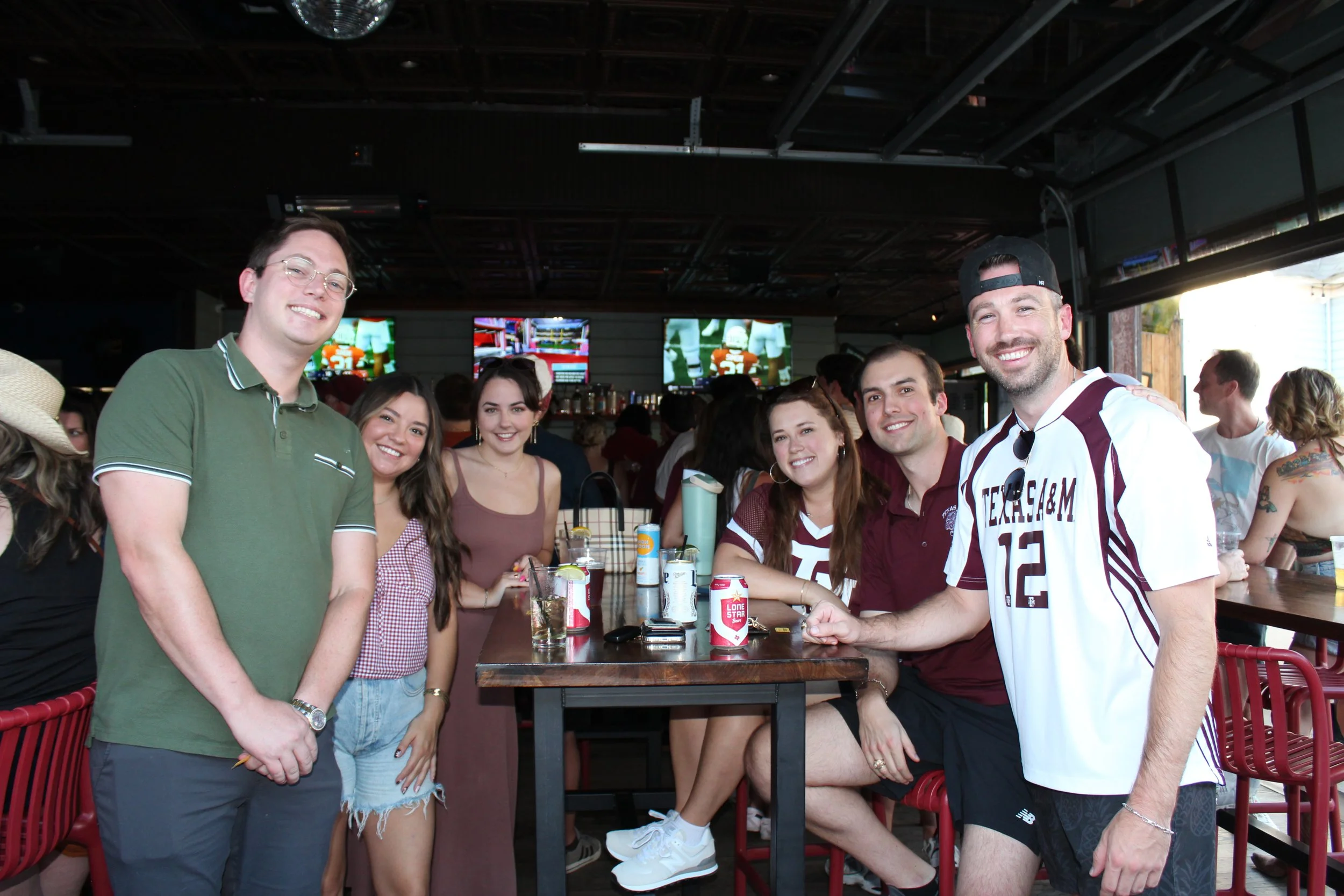 Group of six young people, three men and three women, smiling at camera in a bar or pub. Some are sitting, some standing, with drinks on the table. Background includes bar area and multiple television screens.