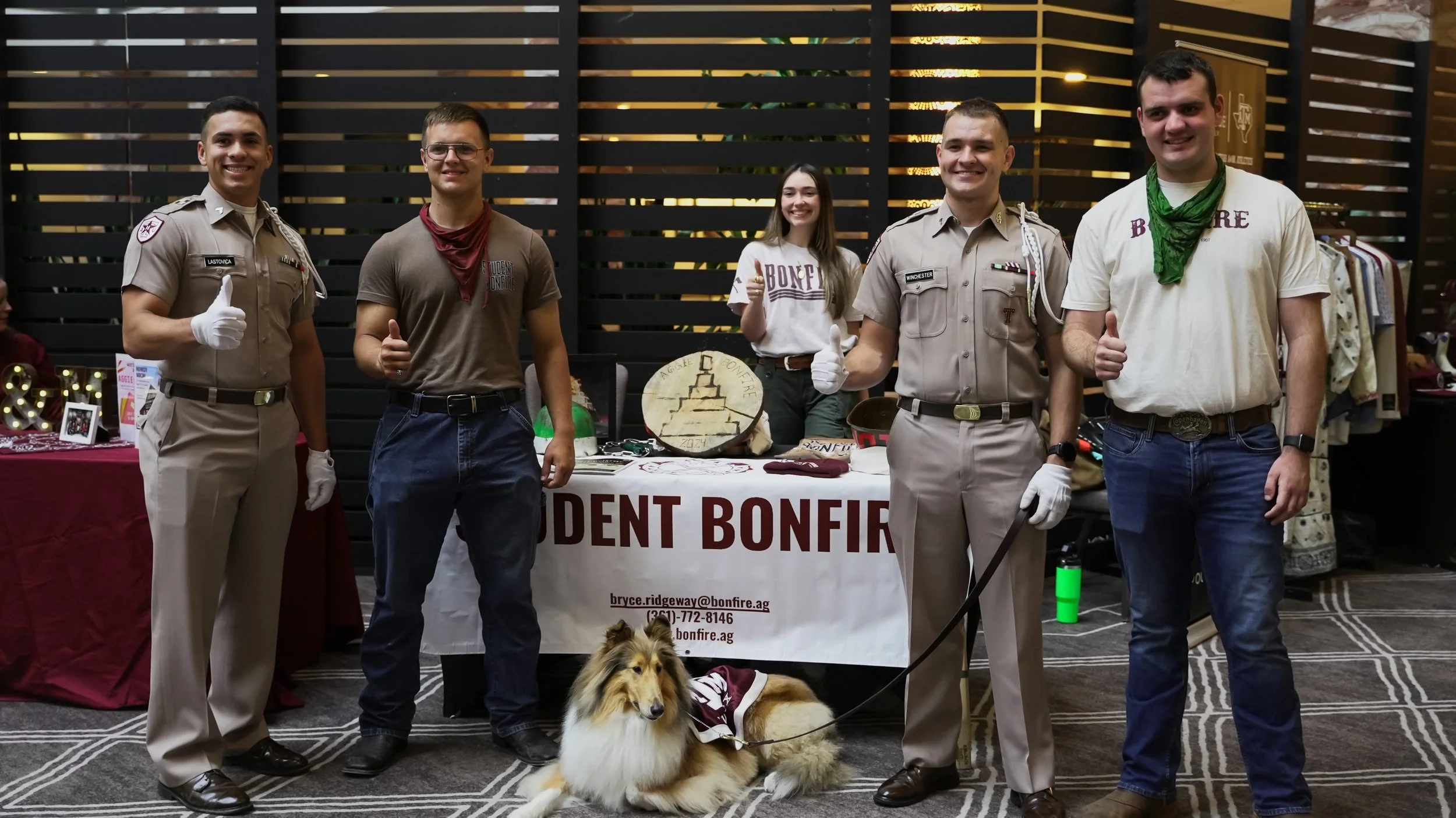 Group of five people, three men and two women, standing behind a table with a sign that reads 'Student Bonfire,' at an indoor event. One man in a military uniform, a dog lying on the floor, and various items on display on the table.