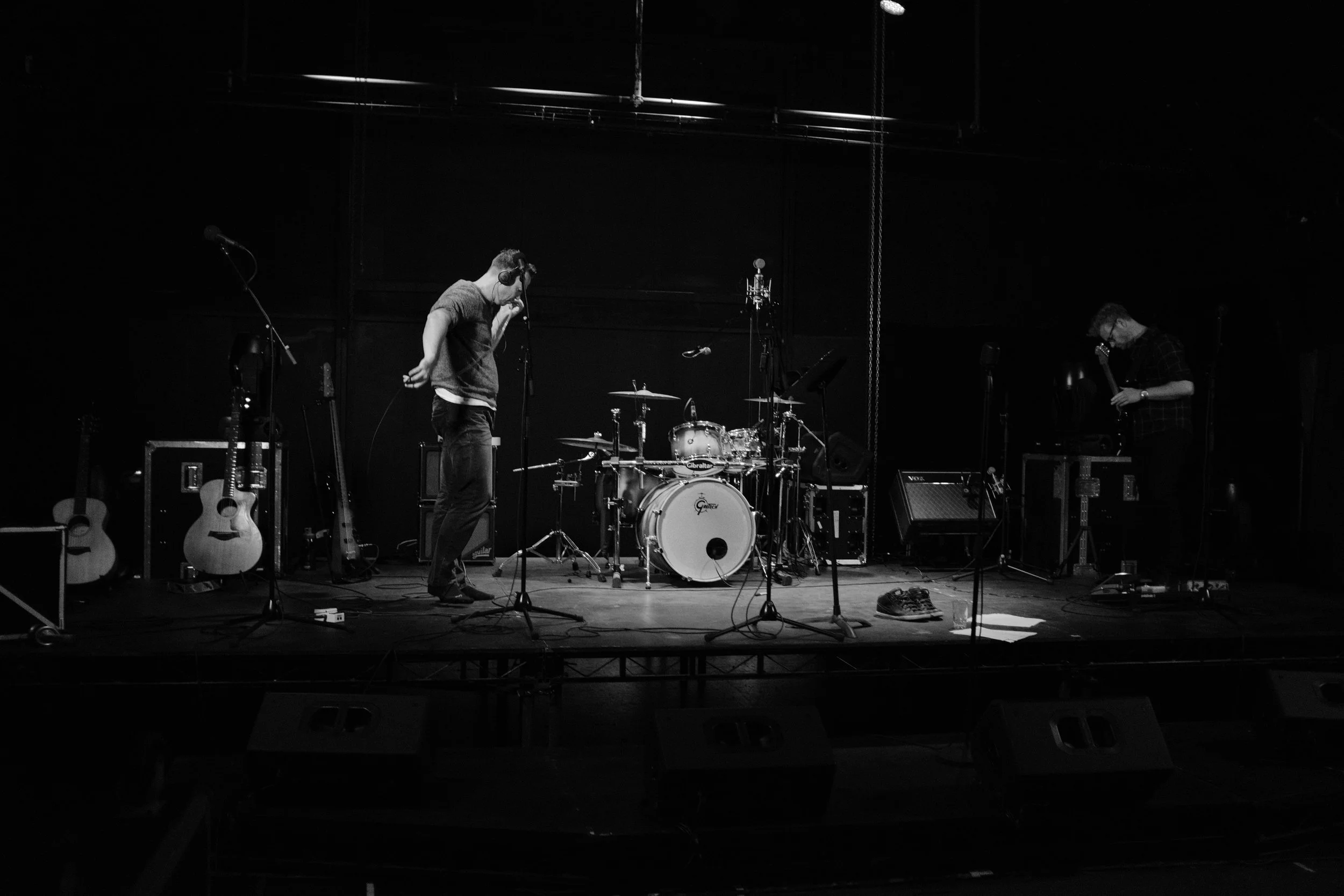 A person standing on a stage in a music venue, preparing for a performance with musical instruments and equipment around, in black and white.