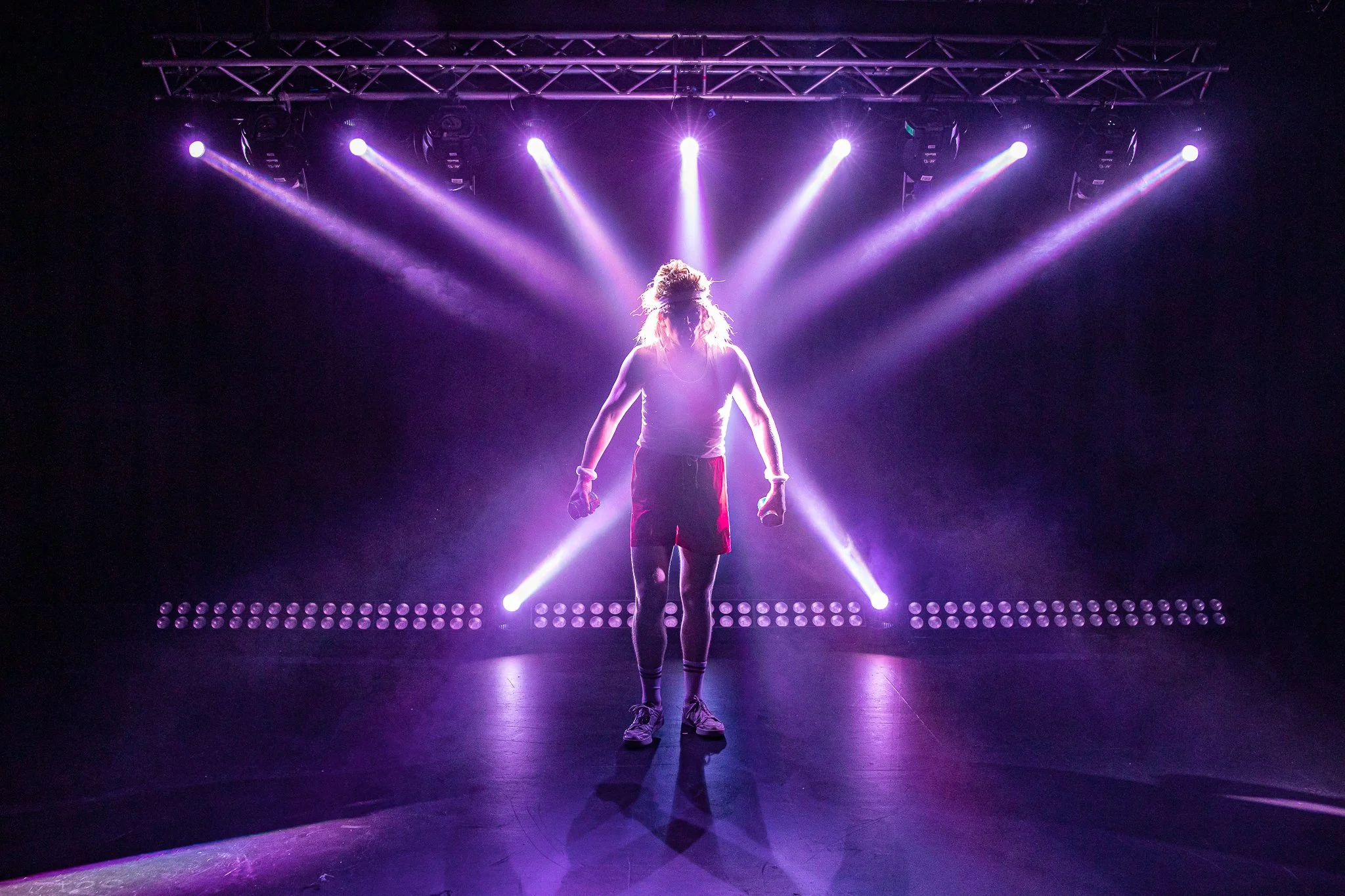 A person standing center stage in front of purple stage lighting, with beams of light radiating from behind, in a dark performance venue.