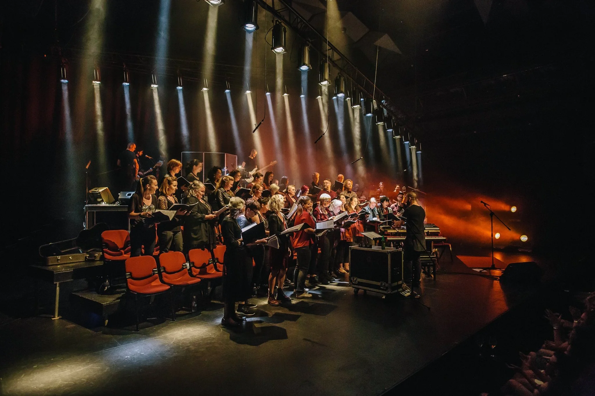 A large choir of men and women performing on stage with conductor, light beams focused on them, and dimly lit auditorium seats in the foreground.