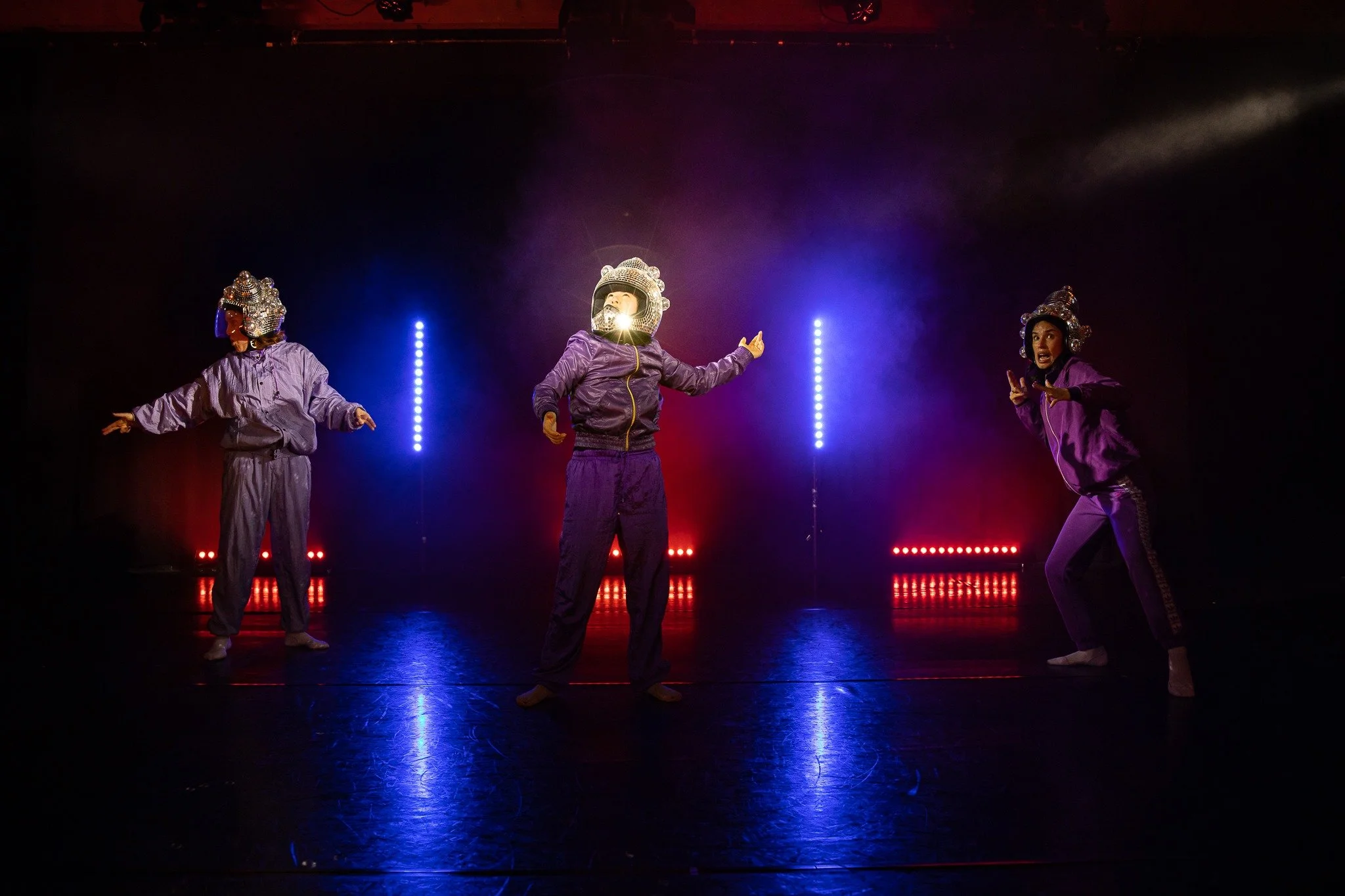 Three performers wearing purple tracksuits and shiny helmets with lights, standing on a stage illuminated by colored lights, engaged in a dance or performance.