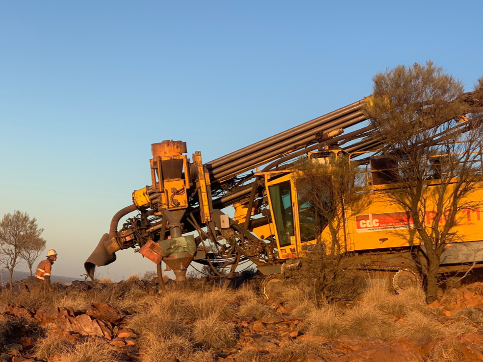 A large yellow drilling machine and a worker in safety gear are working outdoors on a rocky, dry landscape with sparse trees, during sunset or sunrise.